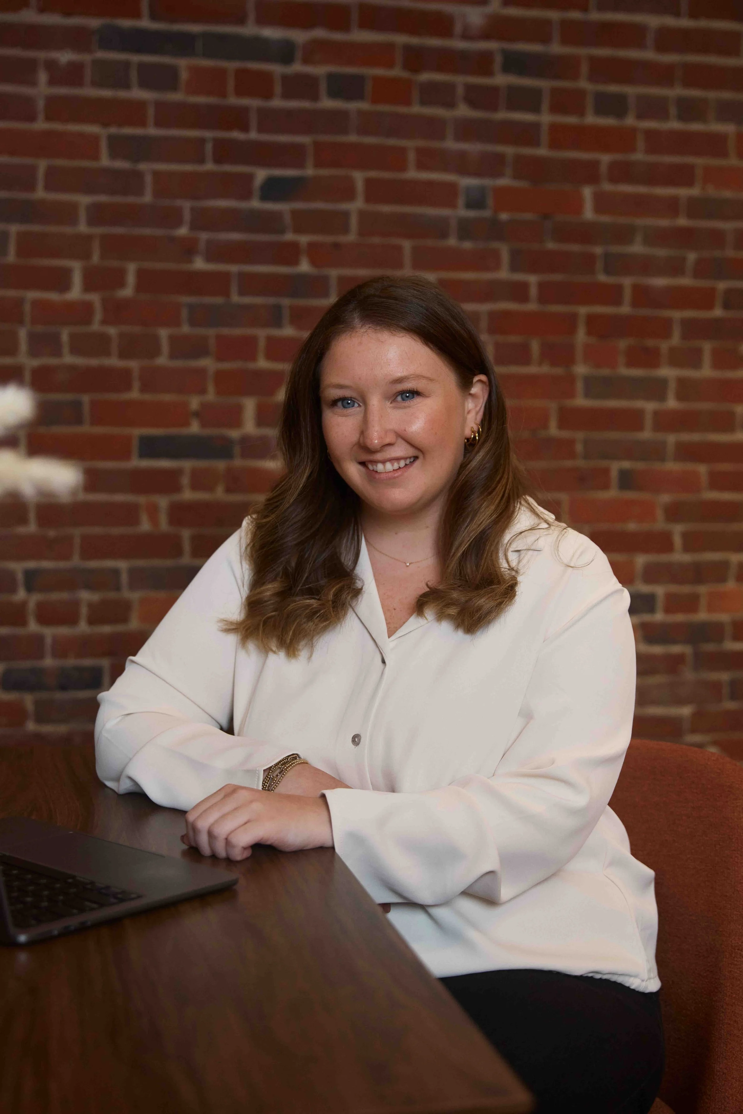 A woman with long brown hair, wearing a white blouse, sitting at a wooden table with a laptop in front of her, smiling against a brick wall background.