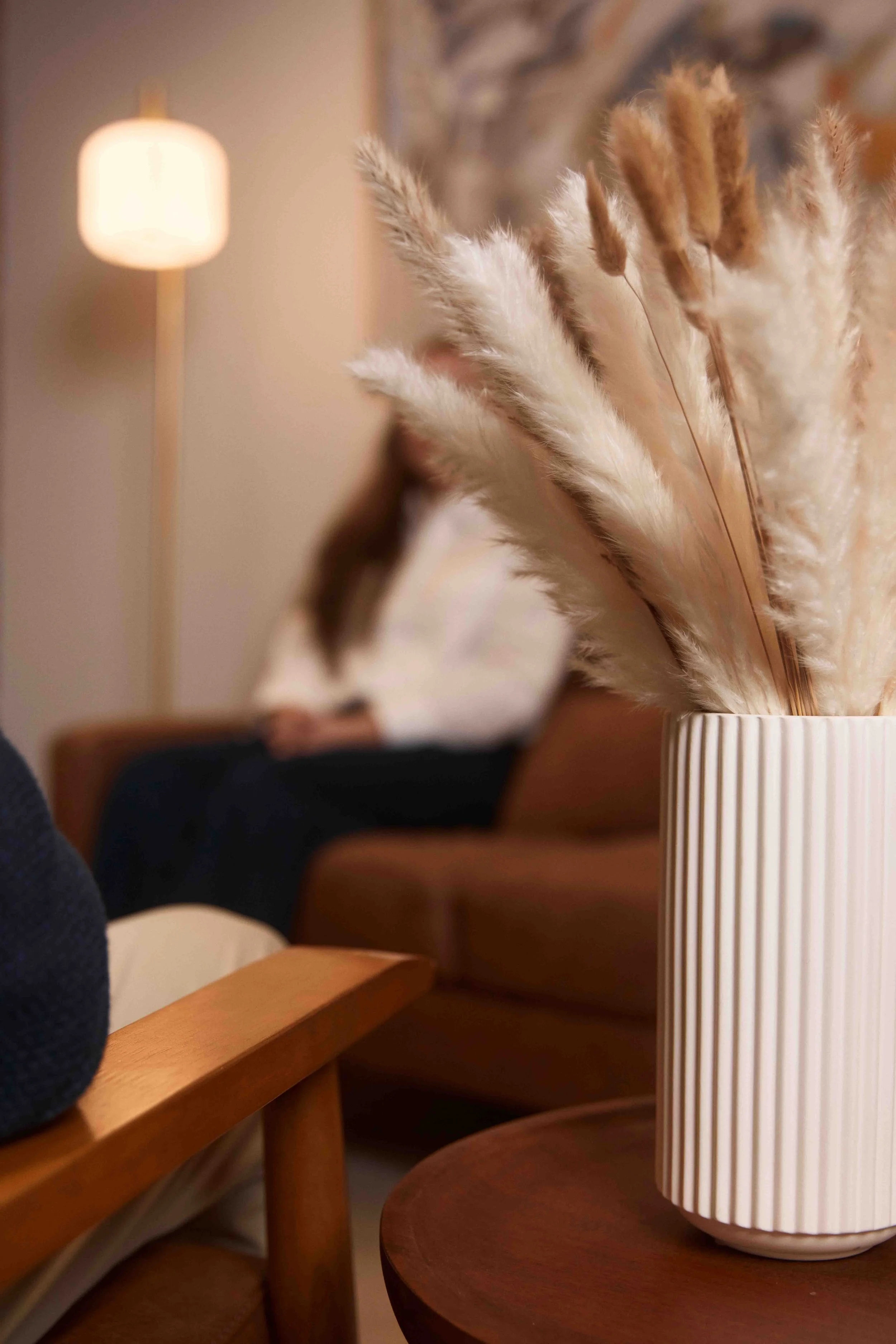 Close-up of a pattern-striped white ceramic vase with dried pampas grass inside, set on a round wooden table in a cozy, softly lit living room. A woman is blurred in the background sitting on a sofa.