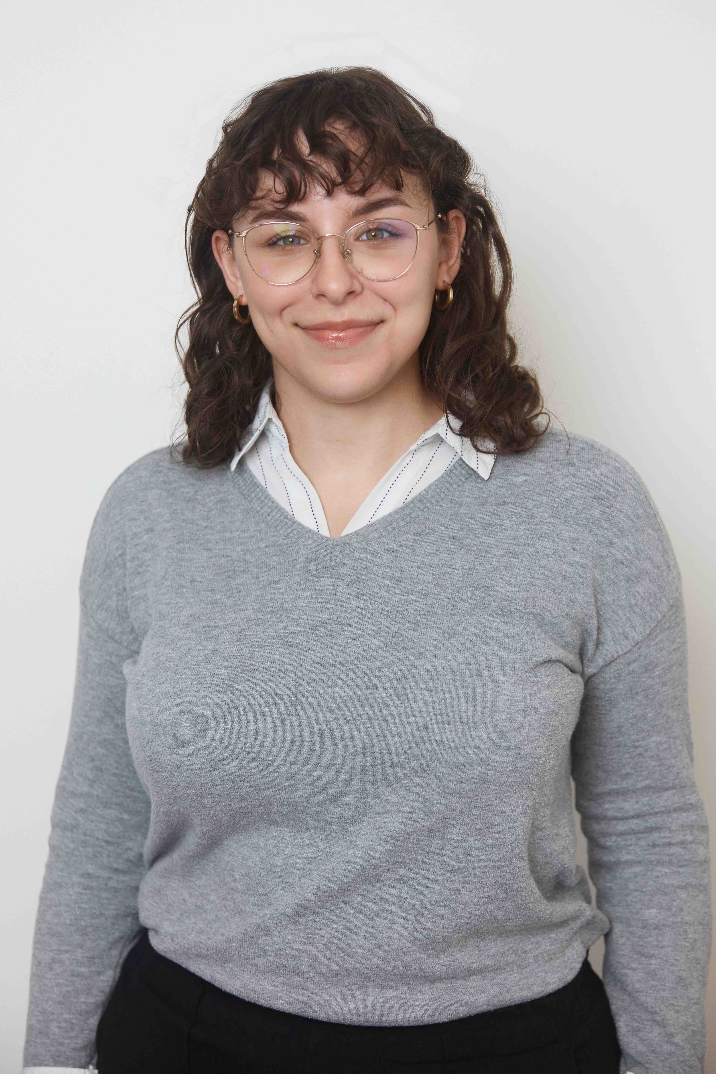 Portrait of a woman with curly brown hair, glasses, gold hoop earrings, wearing a white collared shirt and a gray sweater, smiling against a plain white background.