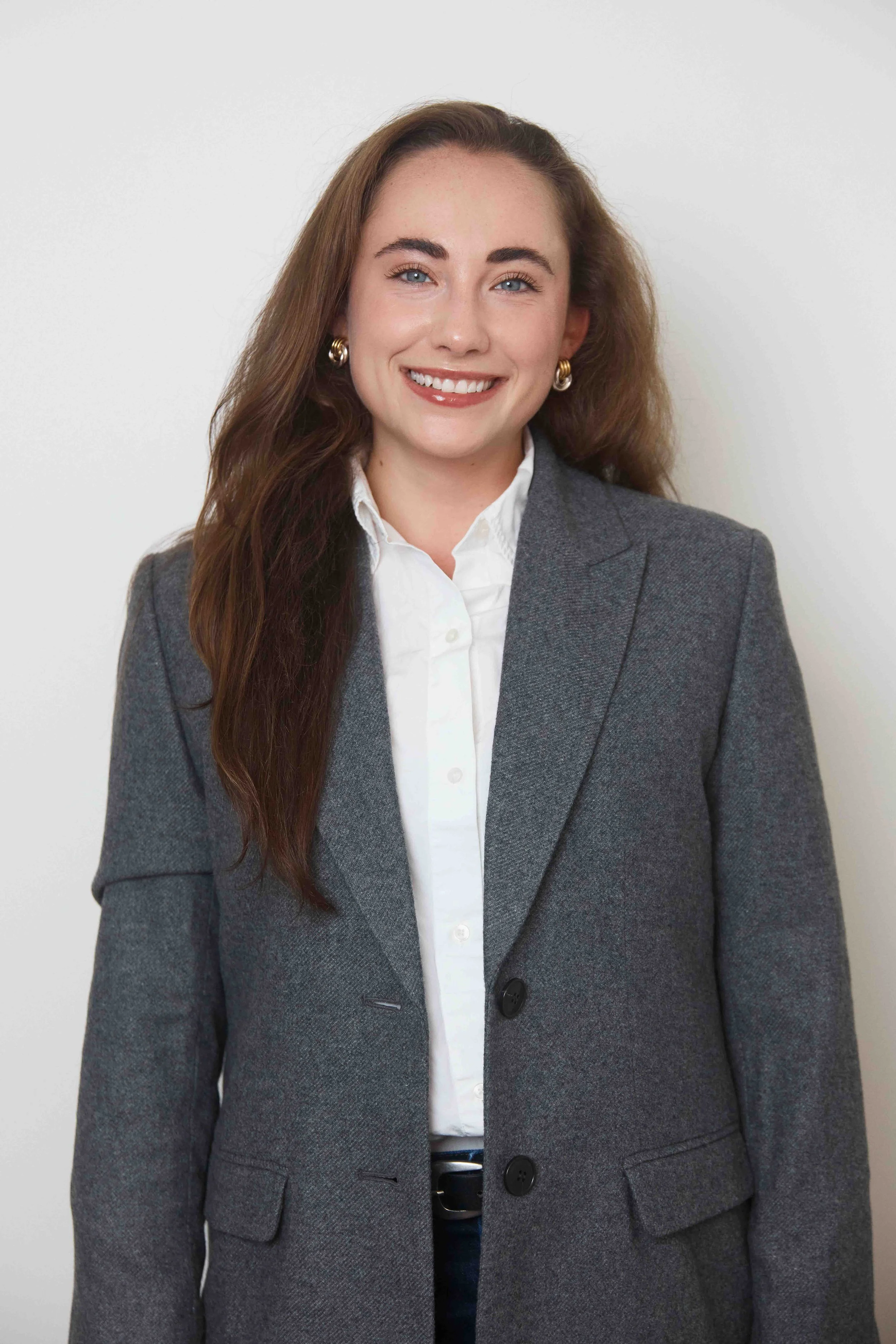 A woman with long brown hair, blue eyes, and light skin, smiling, wearing a grey blazer over a white shirt, with gold earrings, standing against a white background.