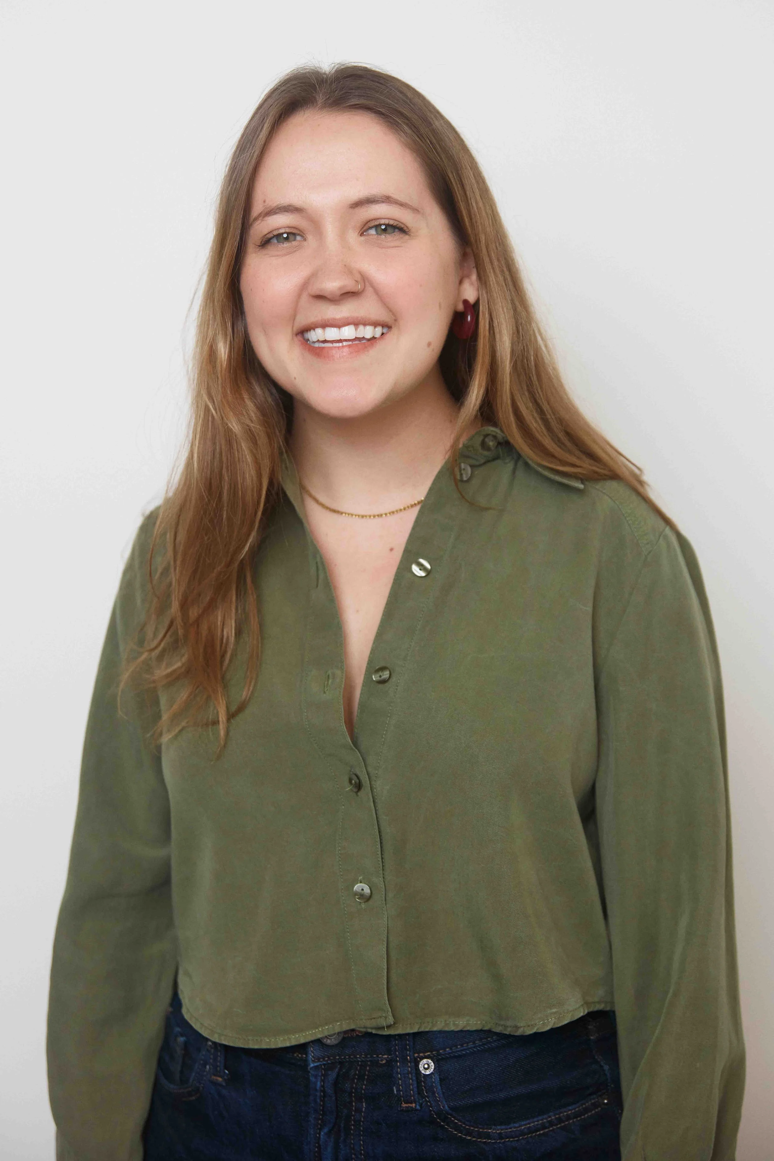 A young woman with long light brown hair, smiling, wearing an olive green button-up shirt and dark jeans, standing against a plain white background.