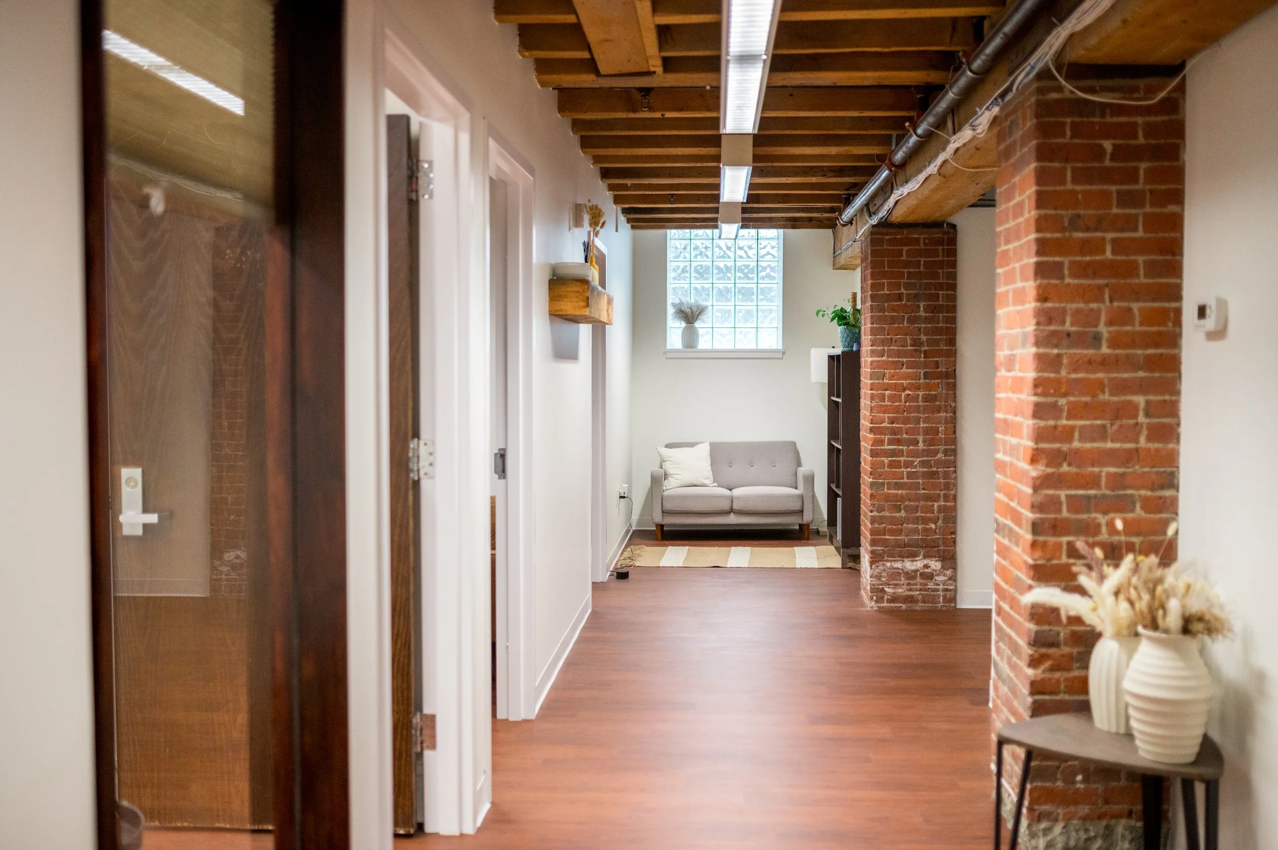 Interior of a modern apartment with exposed brick columns, wooden ceiling beams, a small grey sofa, a window with frosted glass blocks, and decorative elements like vases and plants.