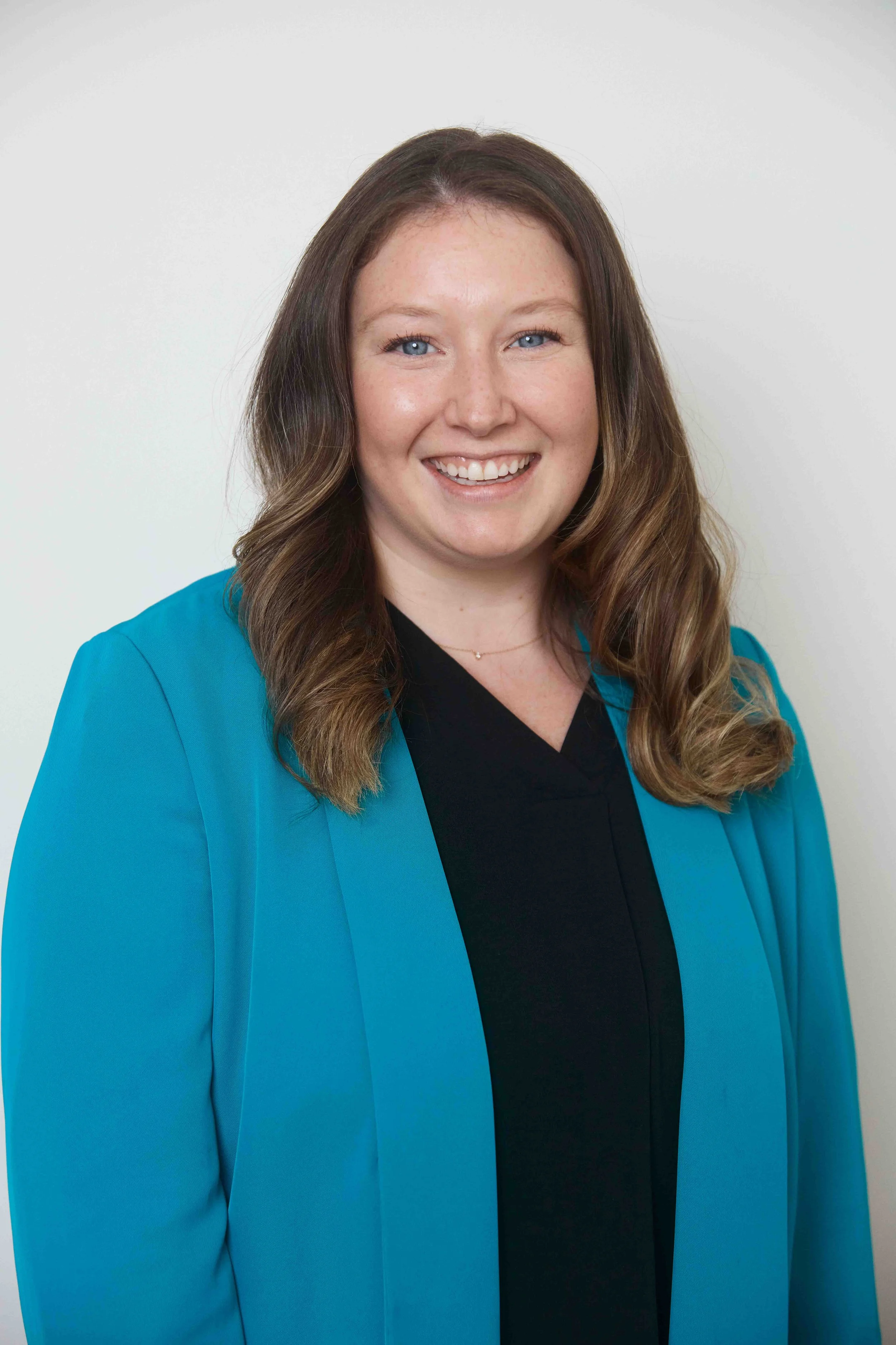 Portrait of a smiling woman with light skin, blue eyes, and shoulder-length wavy brown hair, wearing a black top and a teal blazer, against a plain white background.