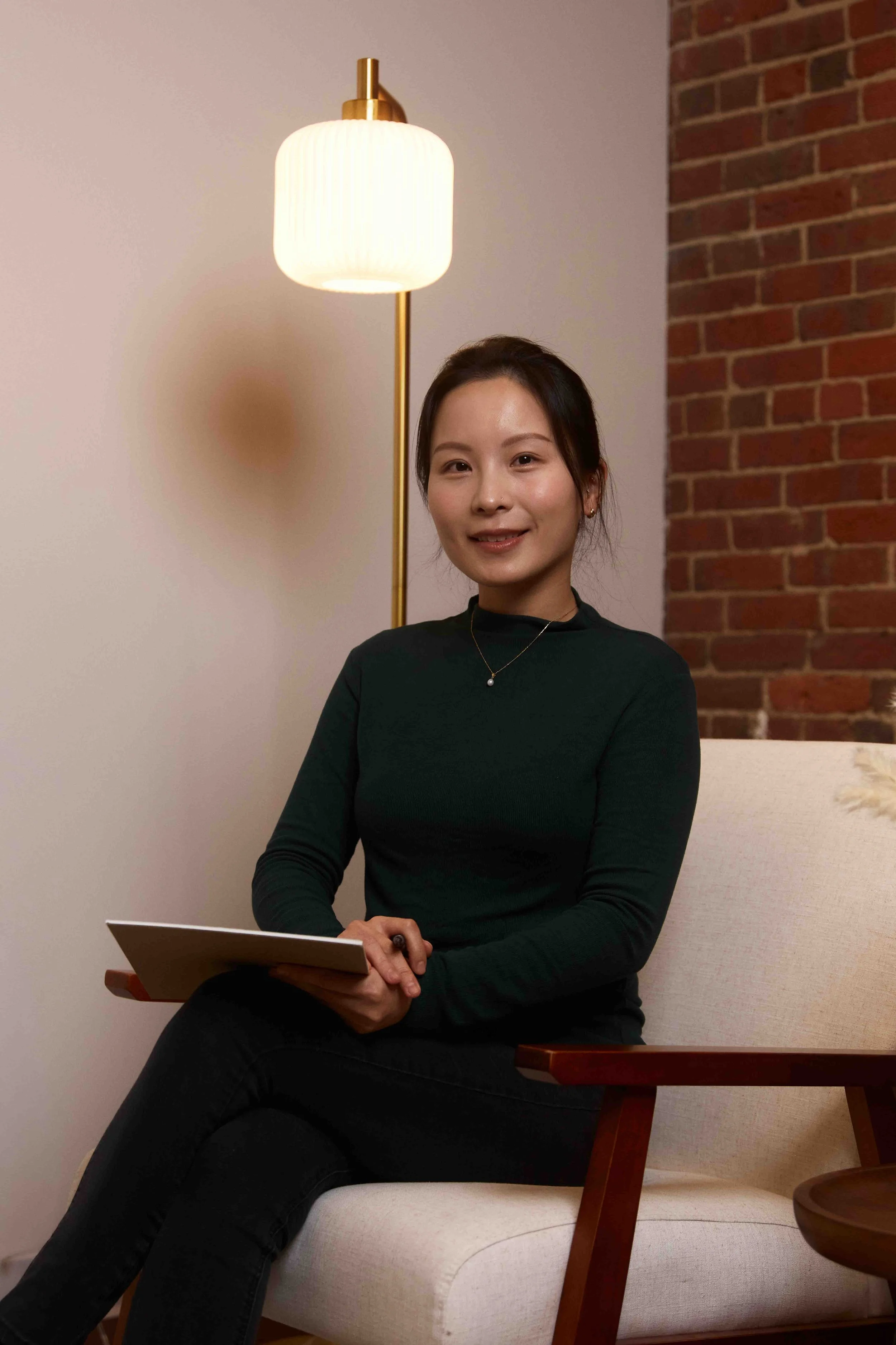 A woman sitting on a white sofa, holding a notebook, smiling in a room with a brick wall and a standing lamp.