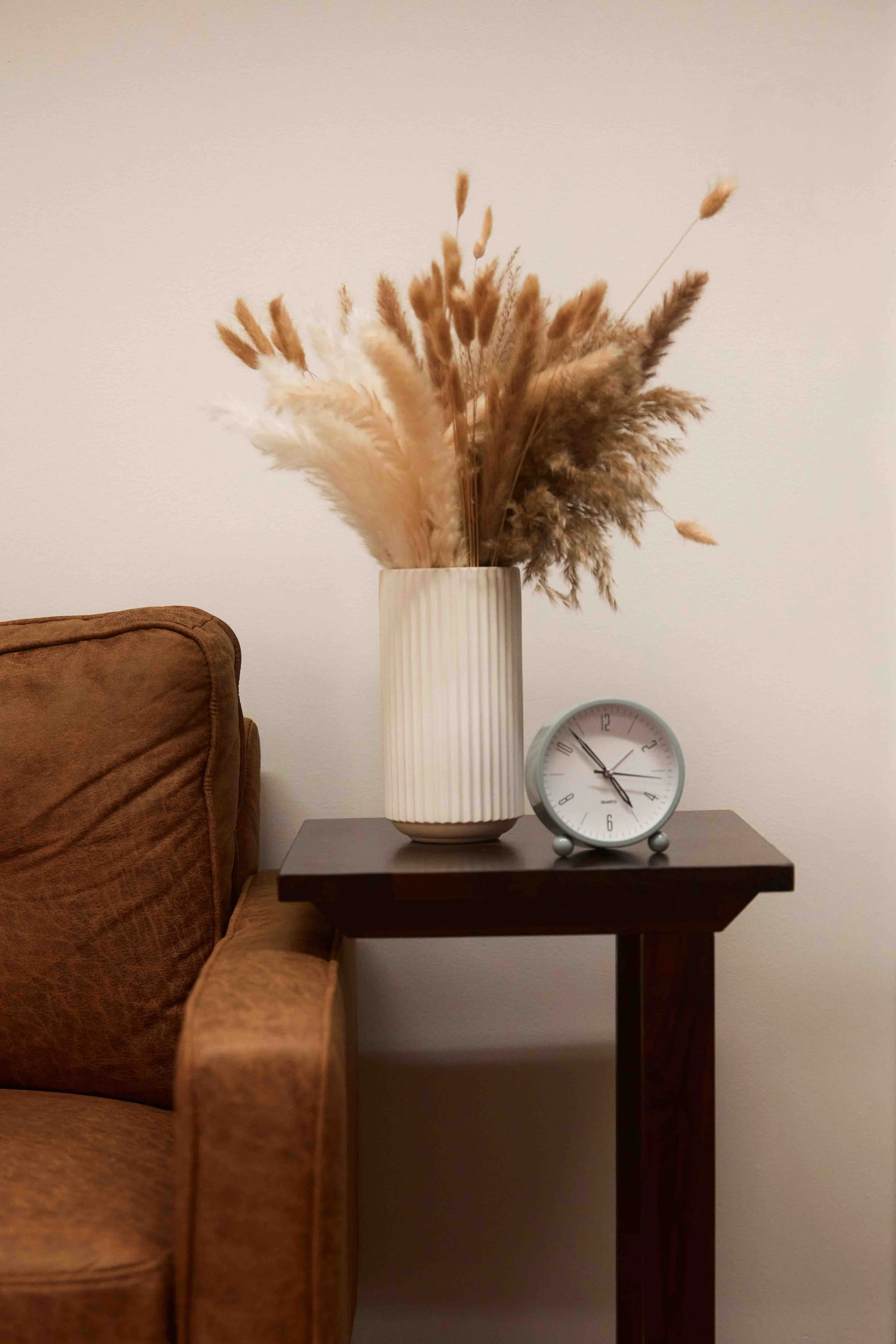 A brown couch next to a dark wood side table with a white vase holding dried beige and brown ornamental grasses and a white clock on the table.