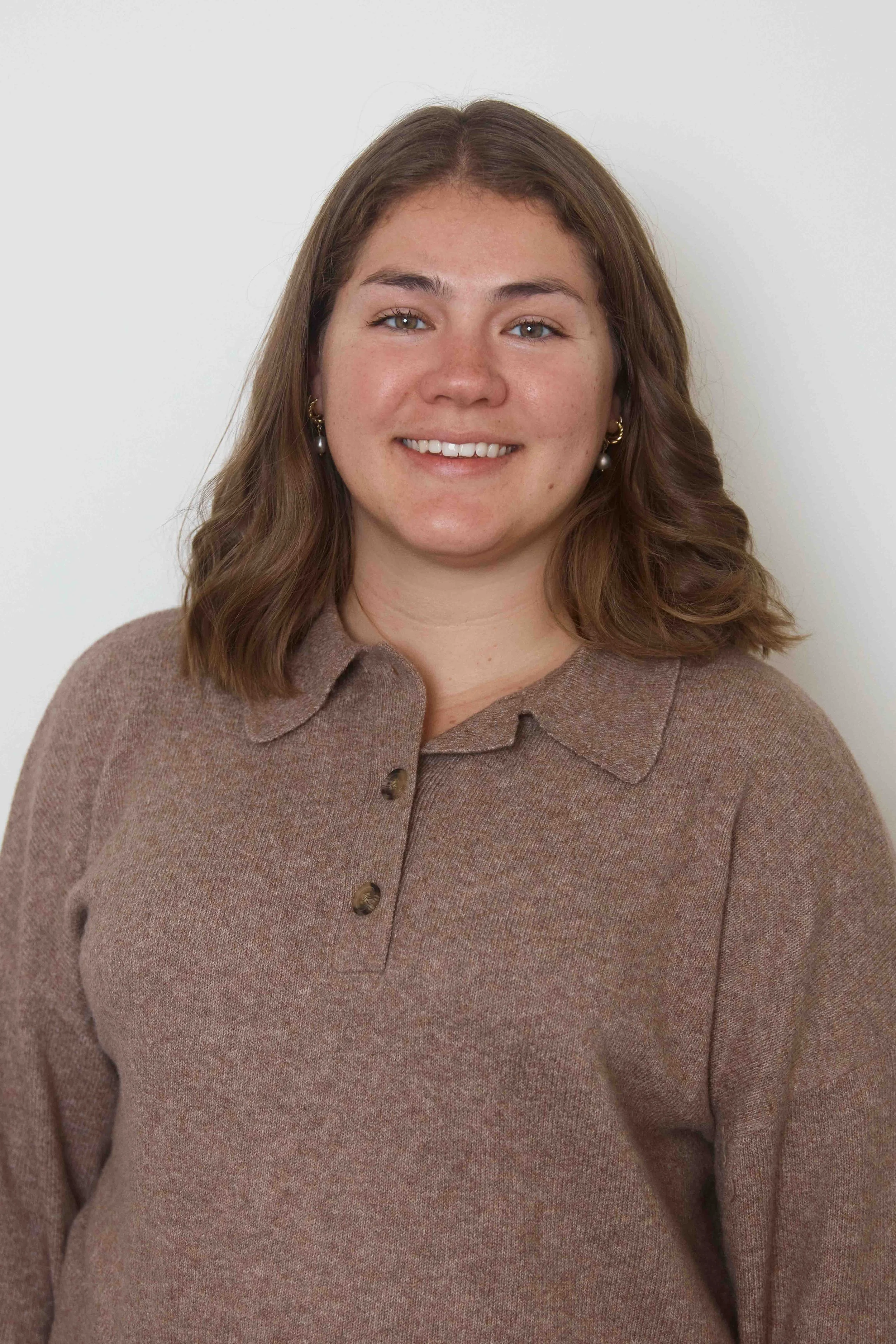 A young woman with shoulder-length brown hair, wearing a brown sweater, smiling and standing against a plain white wall.