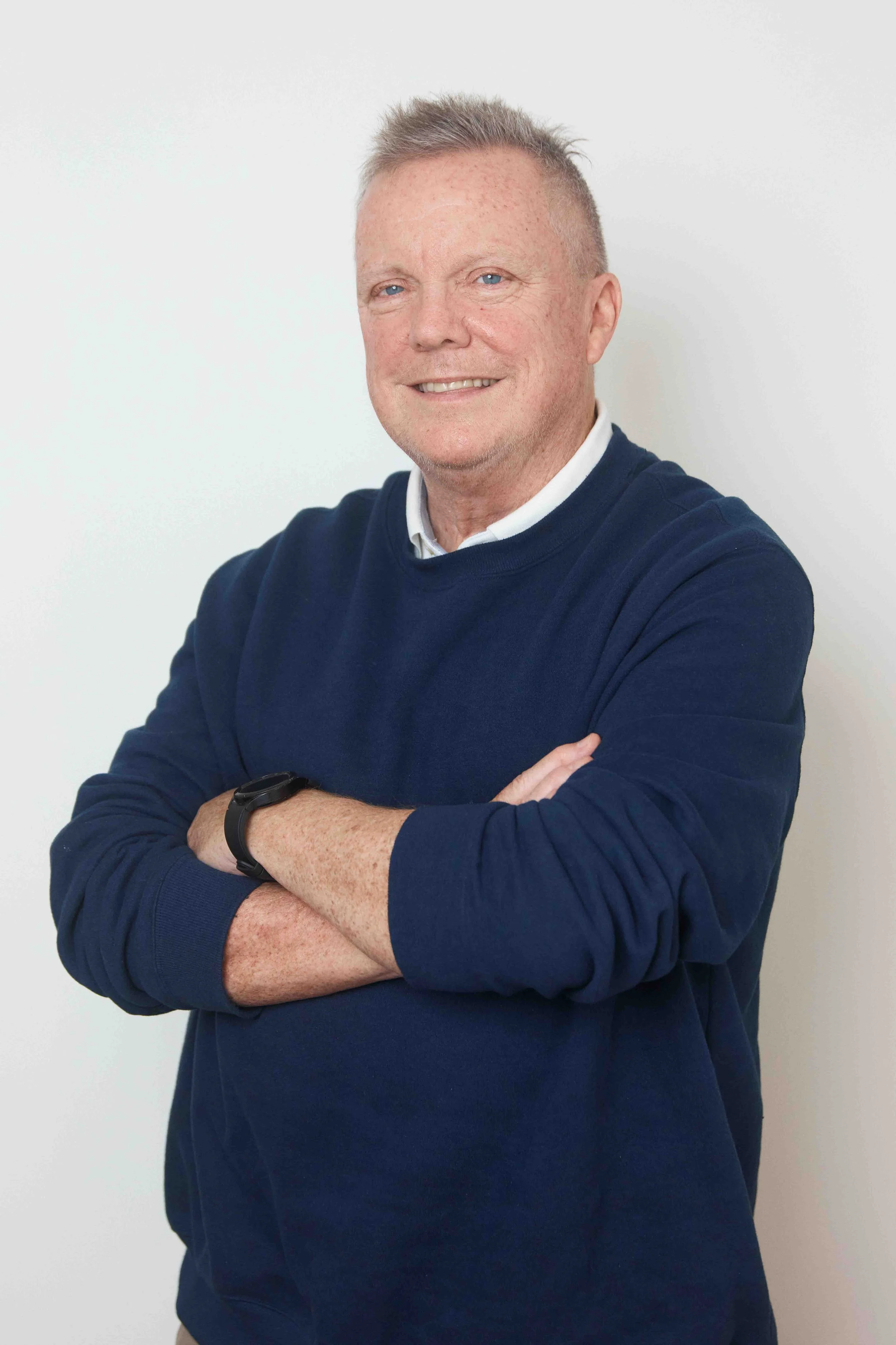 A middle-aged man with short, light-colored hair, wearing a navy blue sweater over a collared shirt, smiling and standing with arms crossed against a plain white background.