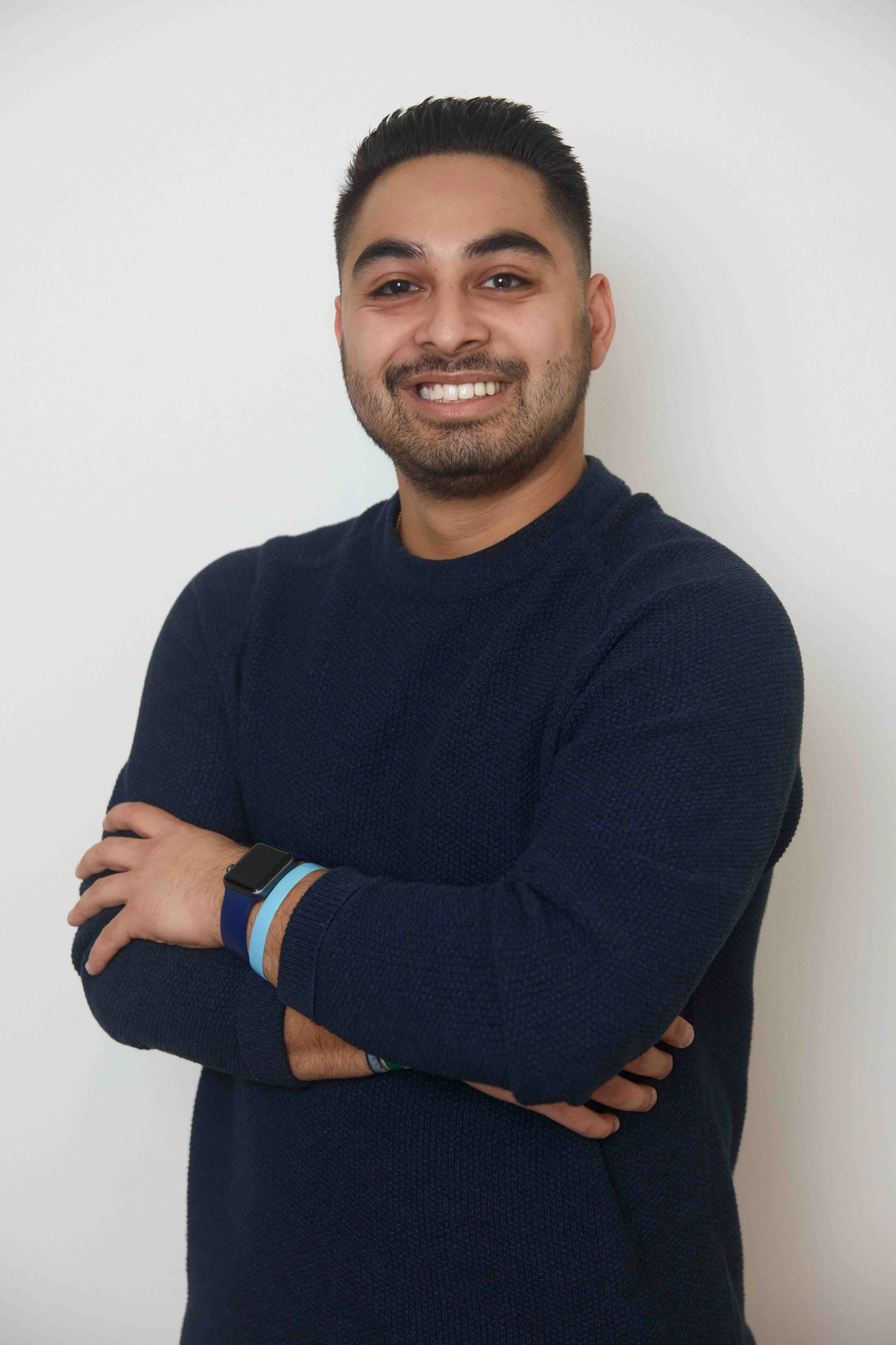 A smiling man with dark hair and a beard wearing a navy blue sweater and a smartwatch, standing against a plain white wall.
