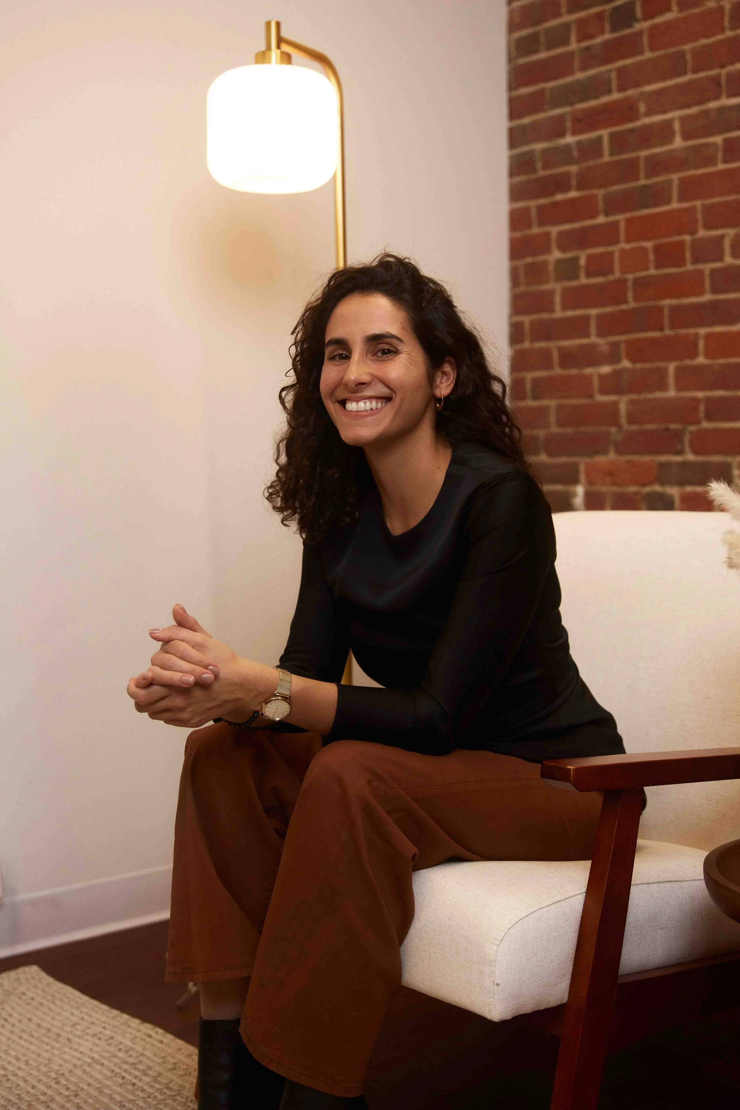 A woman with curly dark hair, wearing a black top and brown pants, sitting on a white cushioned chair, smiling, with a brick wall and a modern wall lamp in the background.