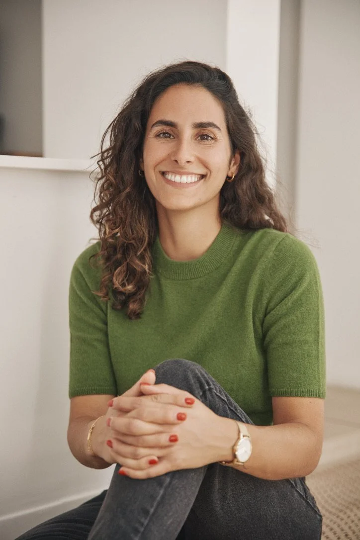 A woman with curly brown hair, wearing a green short-sleeve sweater, sitting on the floor with one leg crossed over the other, smiling at the camera in a bright indoor setting.