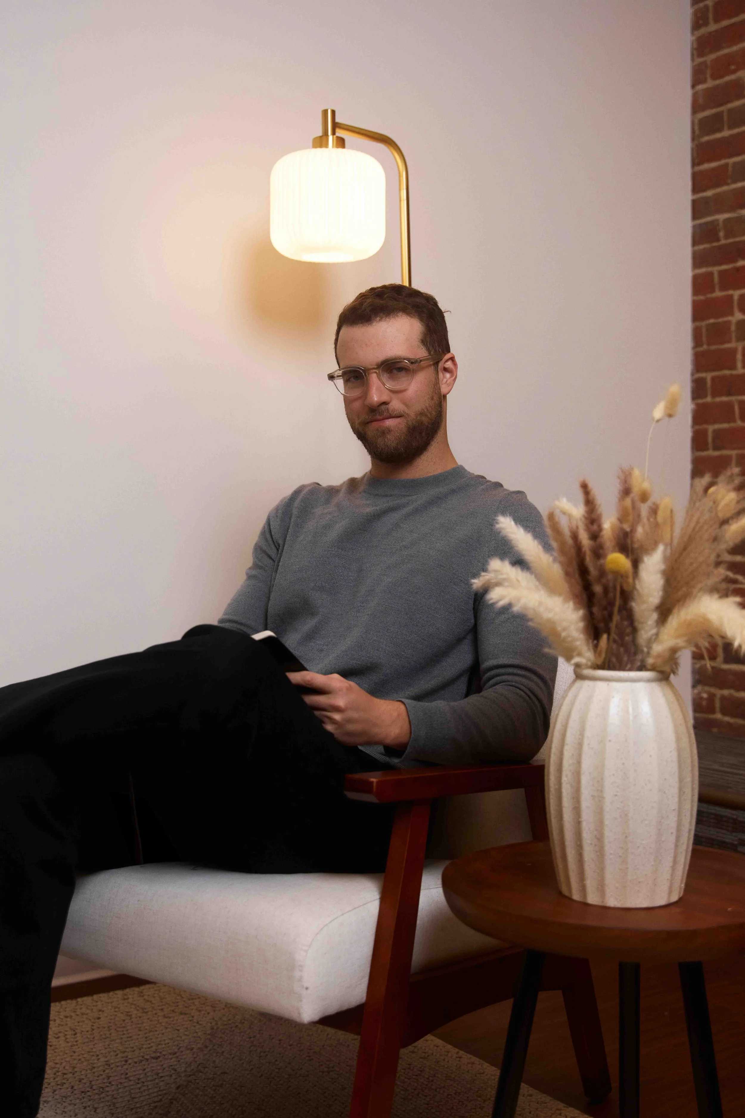 A man with glasses and a beard sitting on a modern beige and wood armchair, holding a tablet, near a table with a vase of dried flowers, in a room with white wall, brick accent wall, and a wall-mounted lamp.