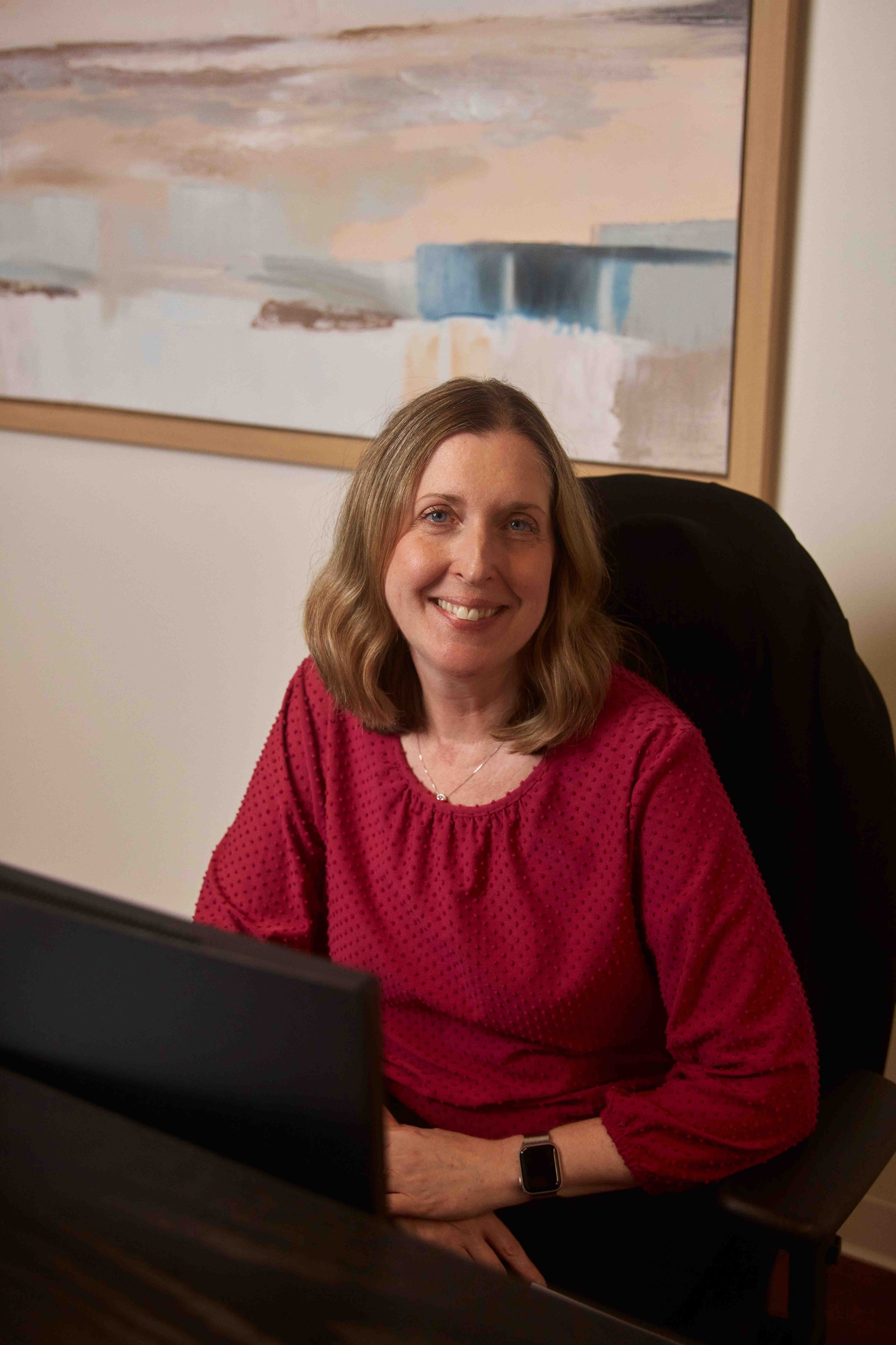 A woman with shoulder-length blonde hair wearing a red blouse, sitting at a desk with a computer monitor, smiling, with a large abstract painting in pastel colors hanging on the wall behind her.