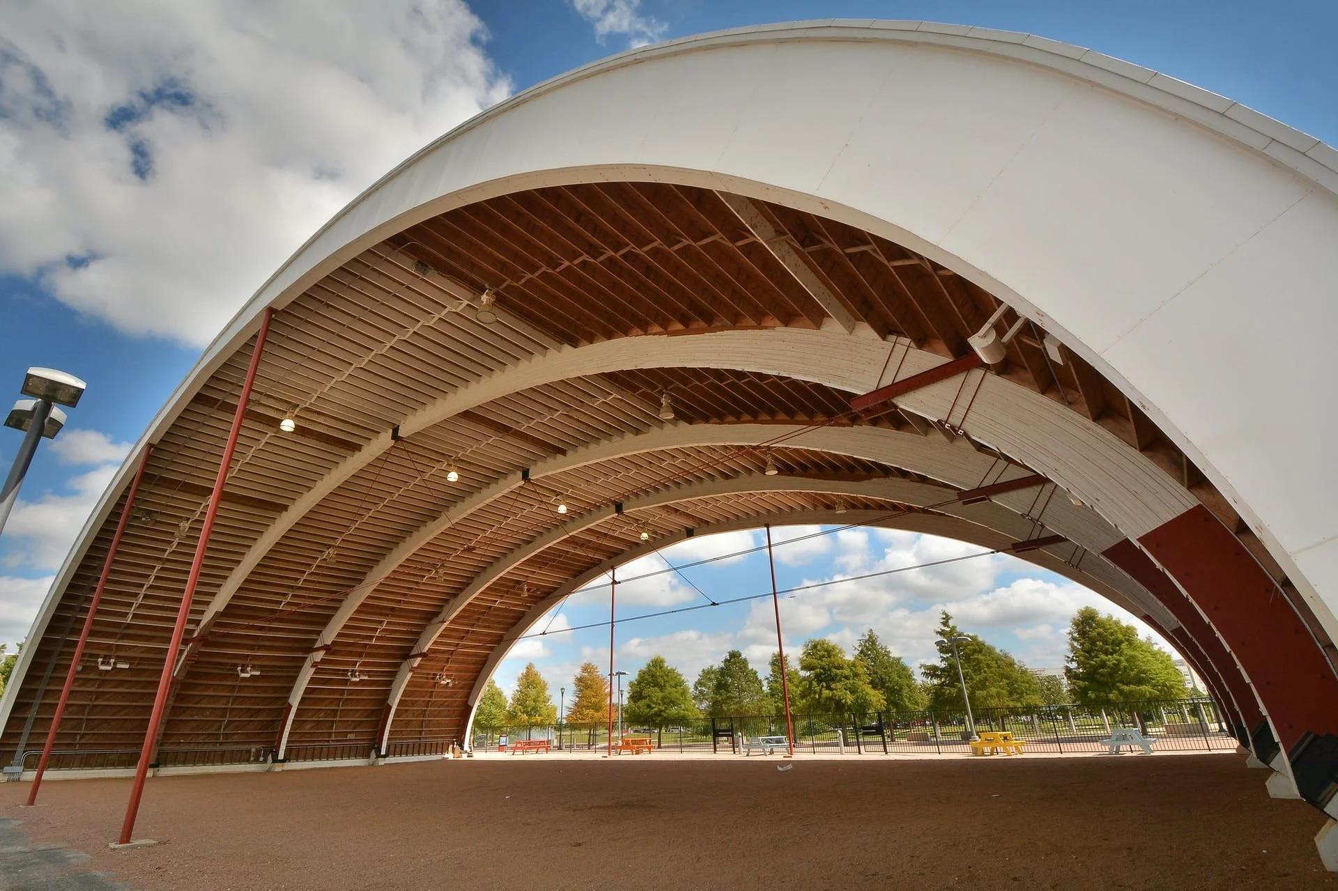 Large open-air arched pavilion with curved wooden beams and a white canopy roof, overlooking a park with trees, benches, and blue sky in the background.