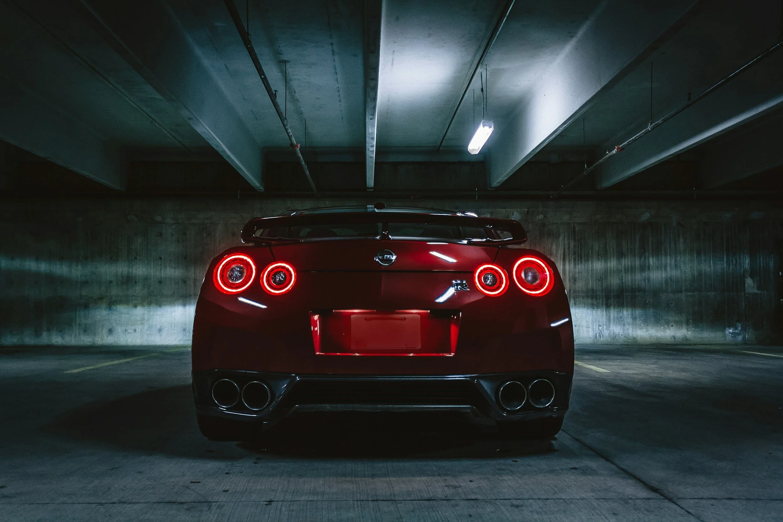 Red Nissan GT-R sports car parked in an underground parking garage, viewed from the rear with illuminated circular taillights and quad exhaust pipes.