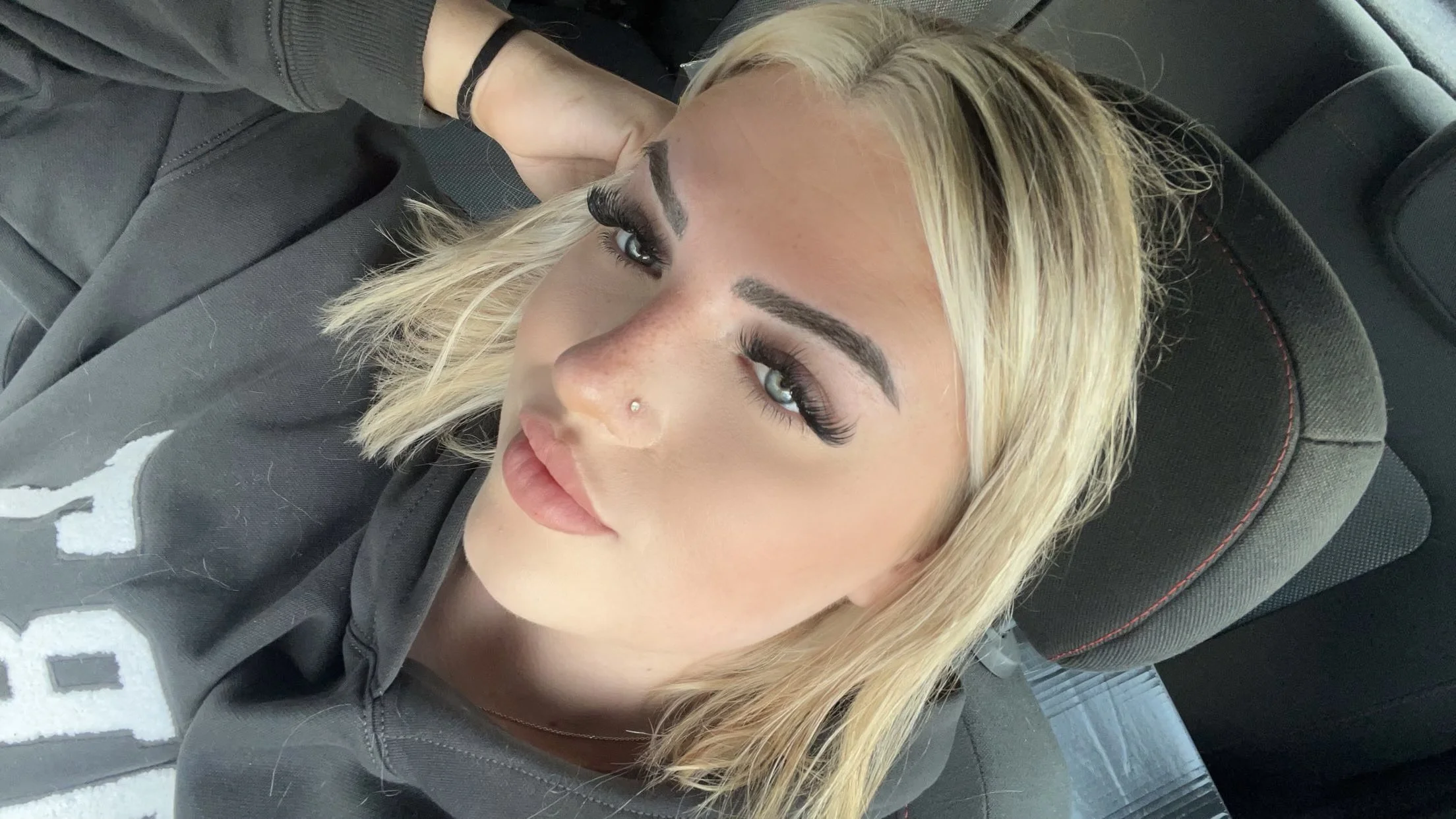 Close-up of a woman with blonde hair and makeup sitting in a car, wearing a dark hoodie, with her hand resting on her head.