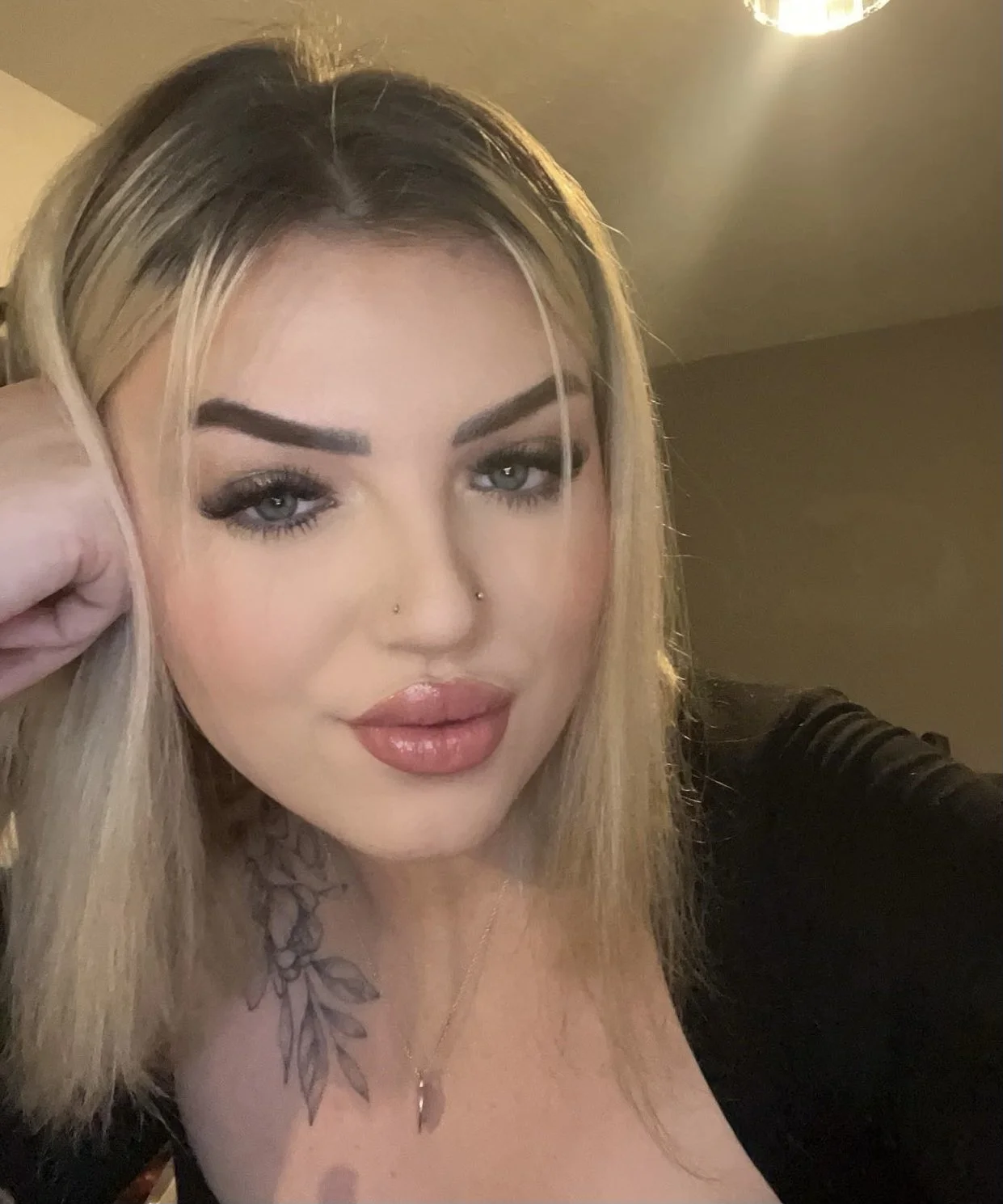 A woman with blonde hair and makeup, wearing a black top and a necklace with a small pendant, posing for a close-up selfie indoors.