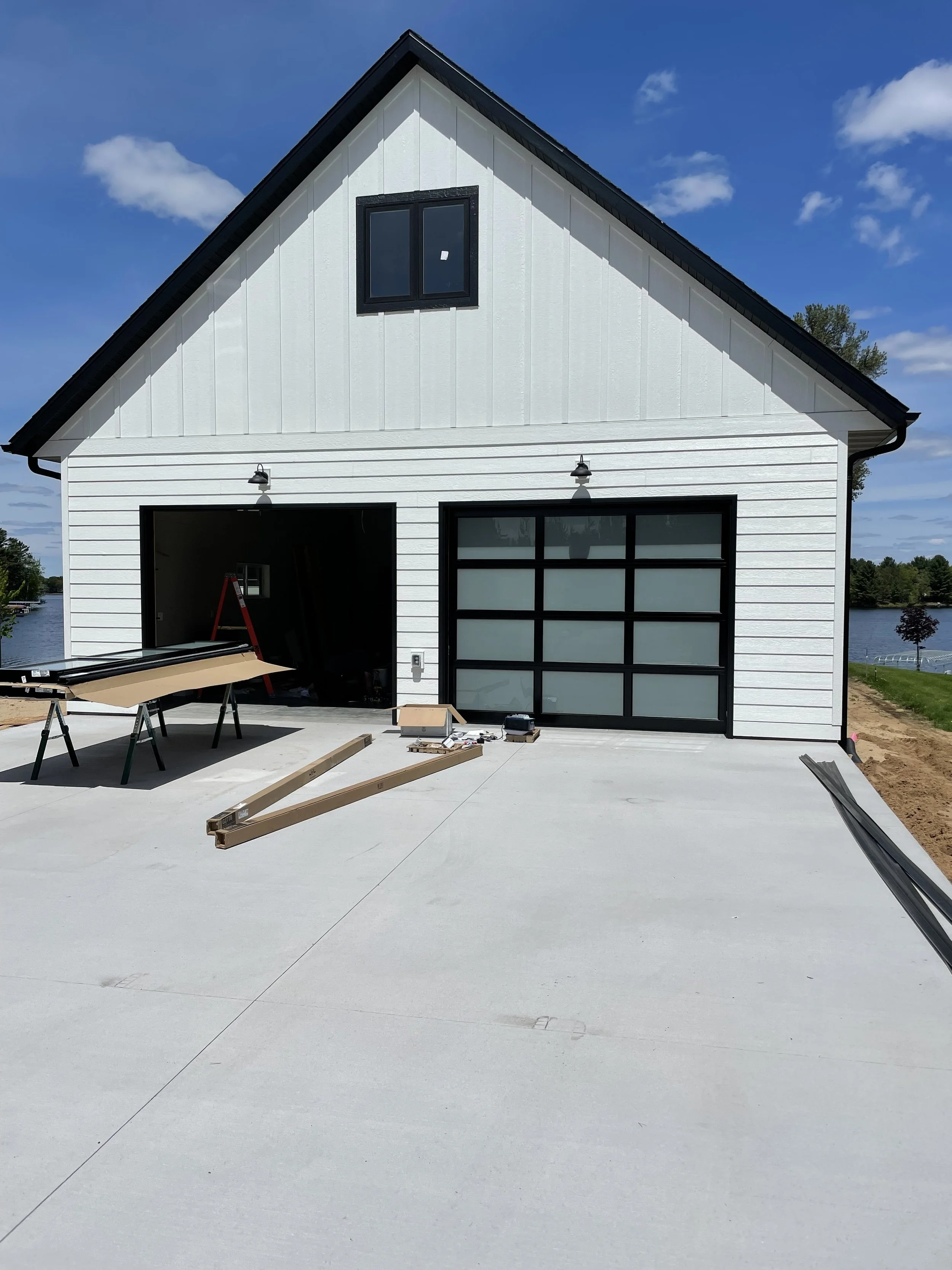 A house under construction with a white exterior, black trim, and a large concrete driveway. The garage door is black with frosted glass panels. Building tools and materials are on the driveway, and a lake and trees are visible in the background.