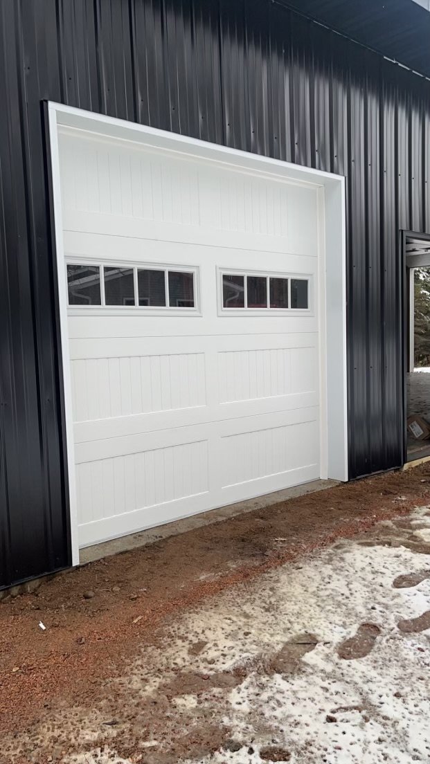 White garage door with small windows on a black metal building with dirt and snow in front.