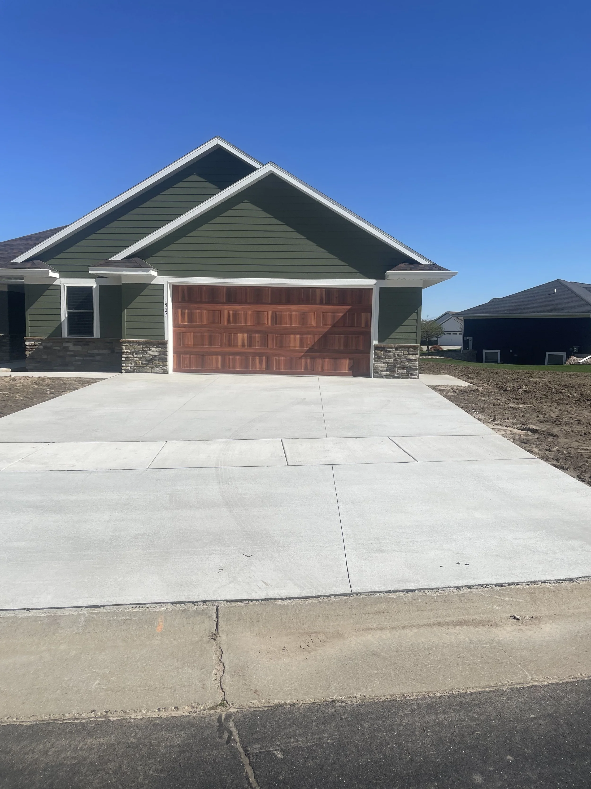 Newly constructed house with a concrete driveway, green siding, stone accents, and a wooden garage door under a bright blue sky.