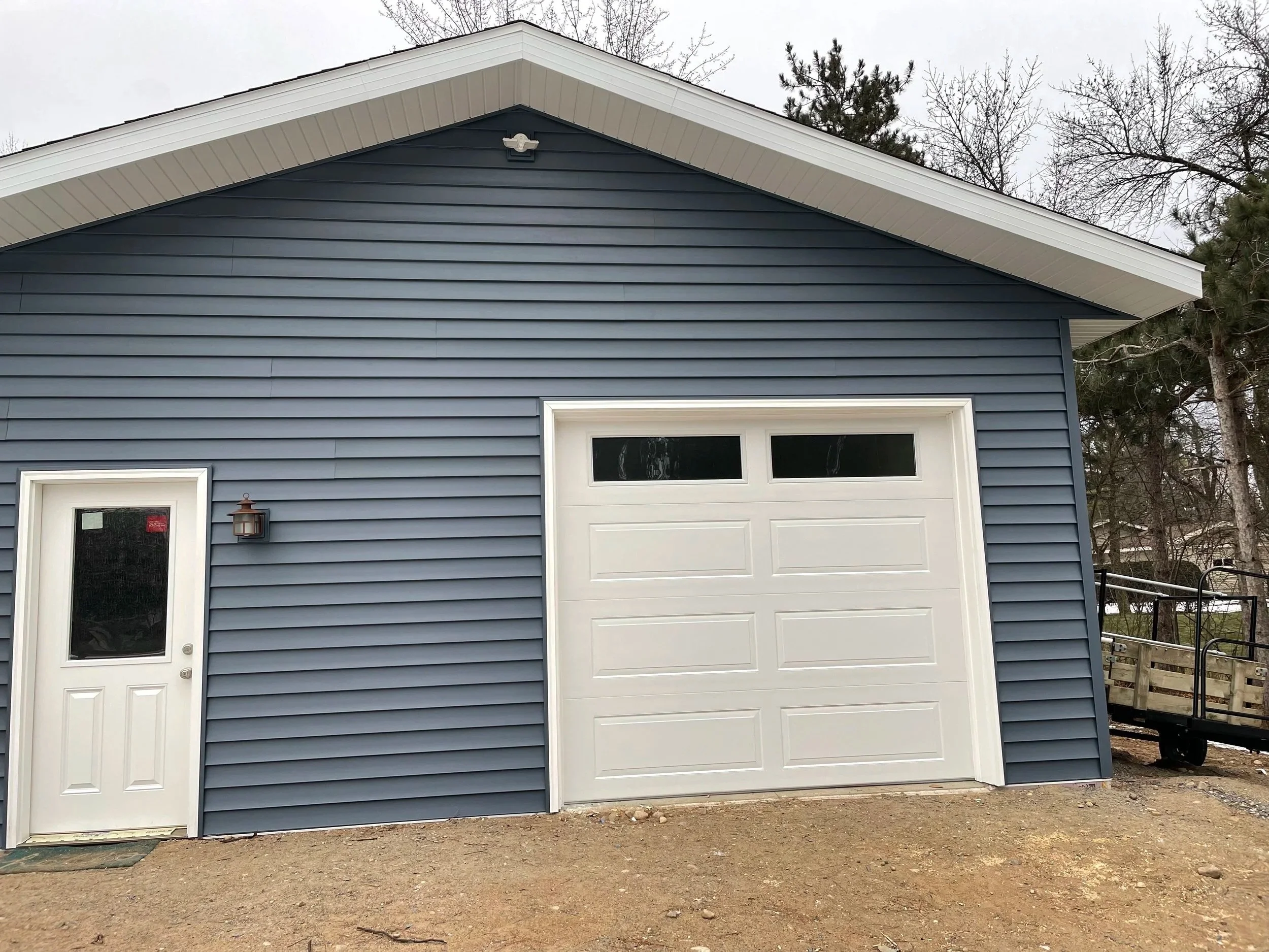 A blue garage with a white roll-up door and a single side door, set on a dirt ground, with leafless trees in the background.