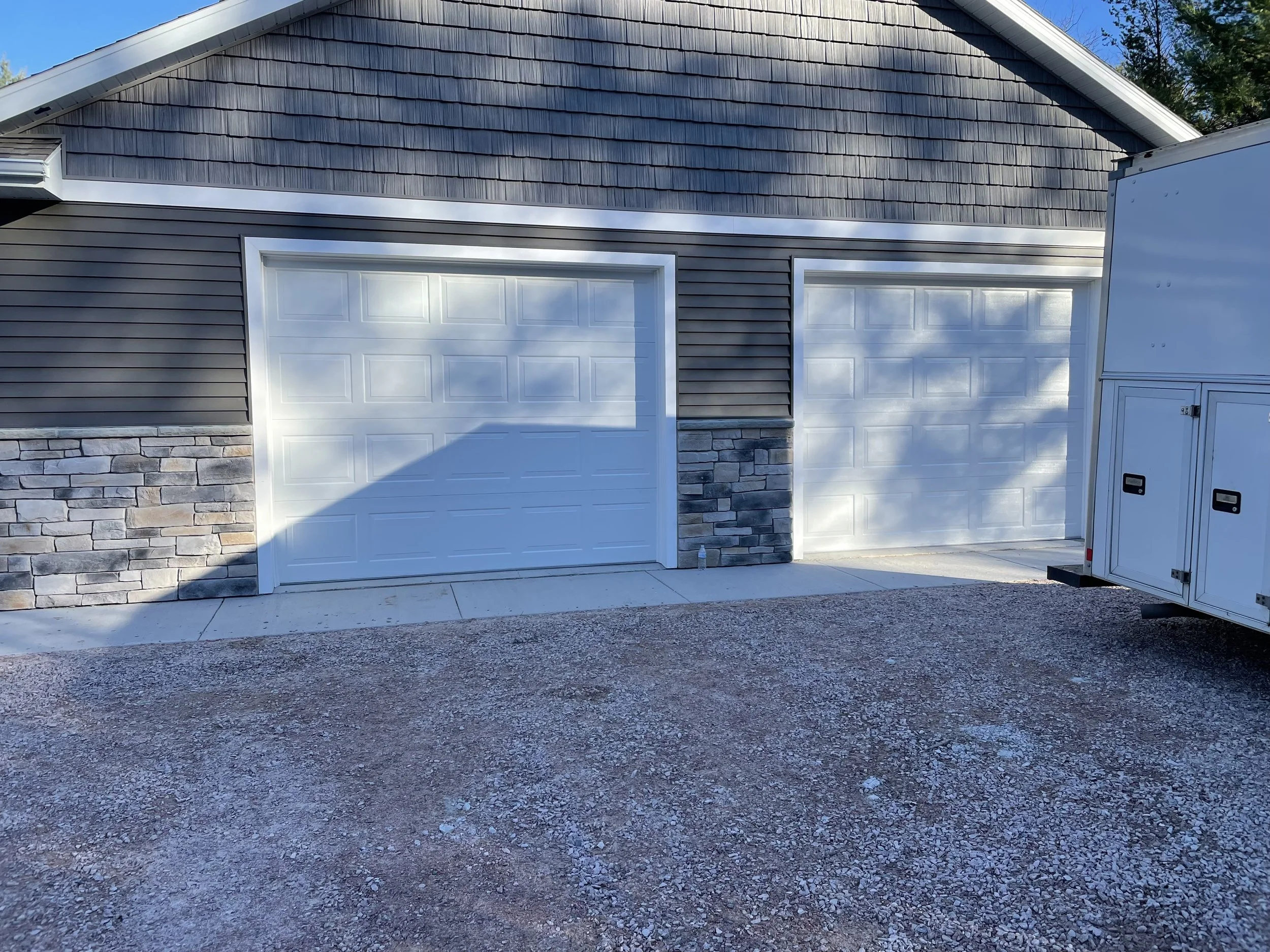 Two white garage doors on a house with dark gray siding, stone accents, and a gravel driveway.