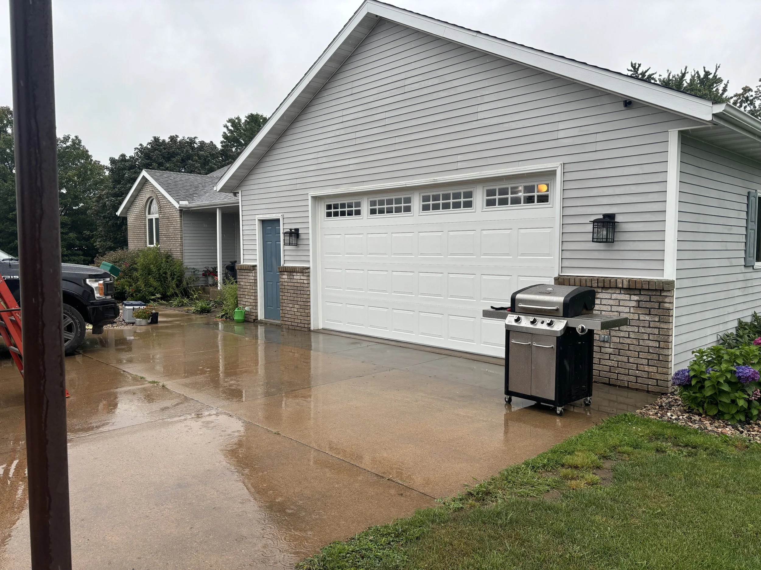 A suburban house with a white garage door, a propane grill, and a driveway wet from rain, with cloudy sky and trees in the background.