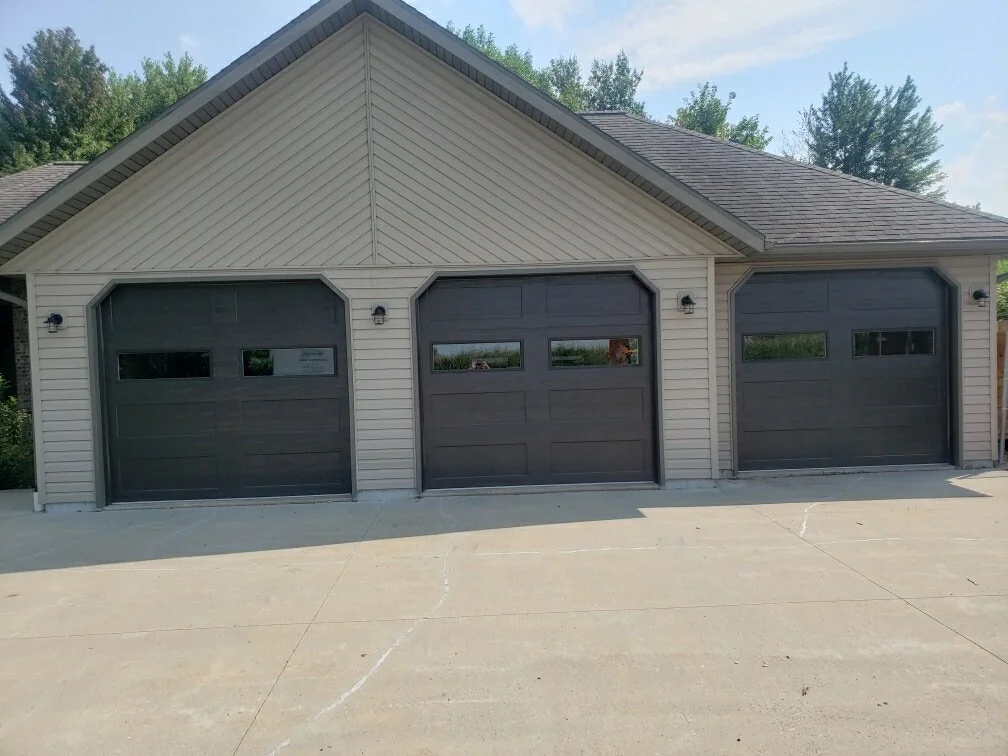 Three black garage doors on a beige house with siding, with trees in the background and a concrete driveway in front.