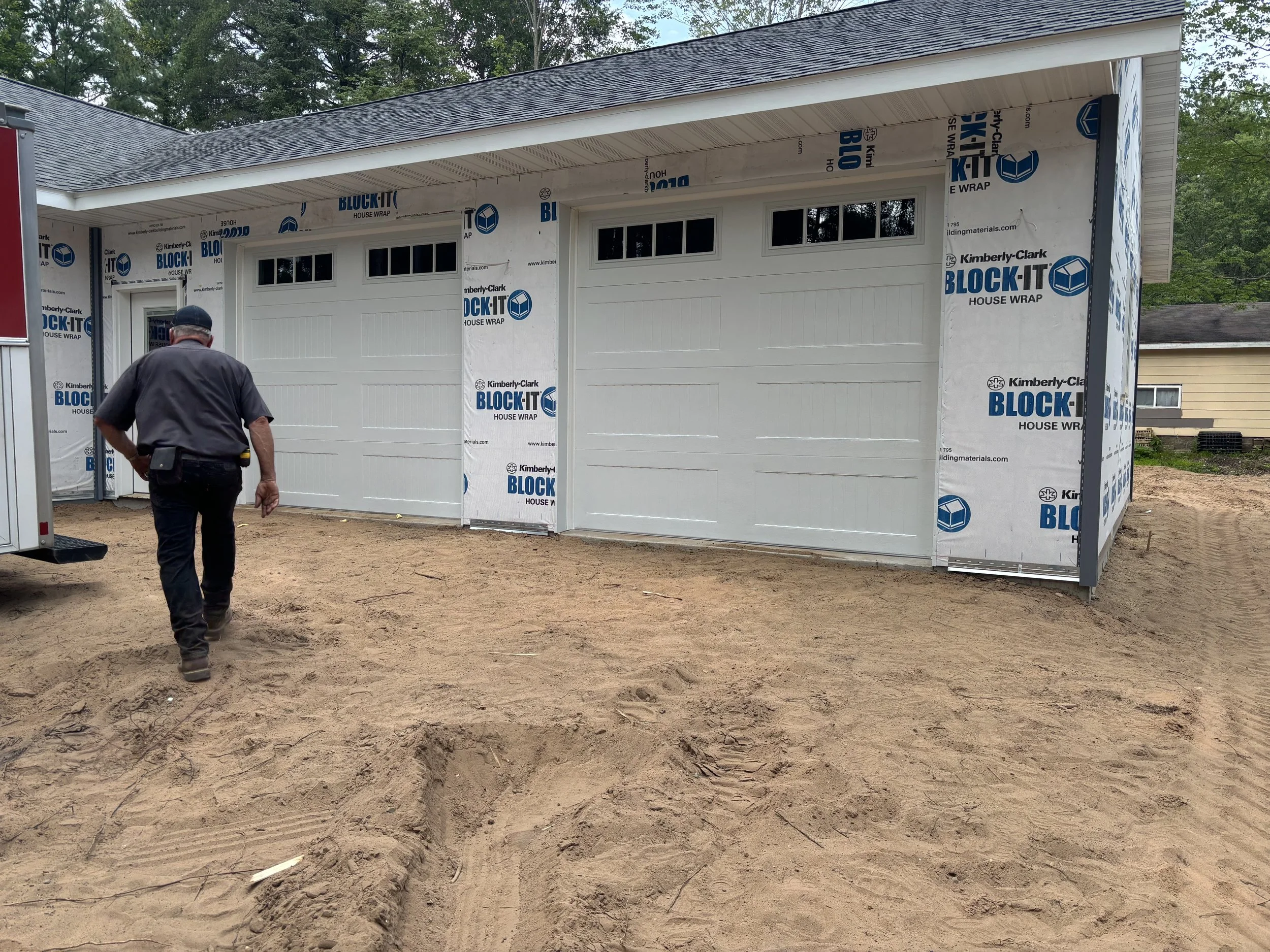 A man walking on a dirt ground in front of a partially constructed garage with white doors, covered with house wrap branded 'Kimberly Clark BLOCK-IT'.