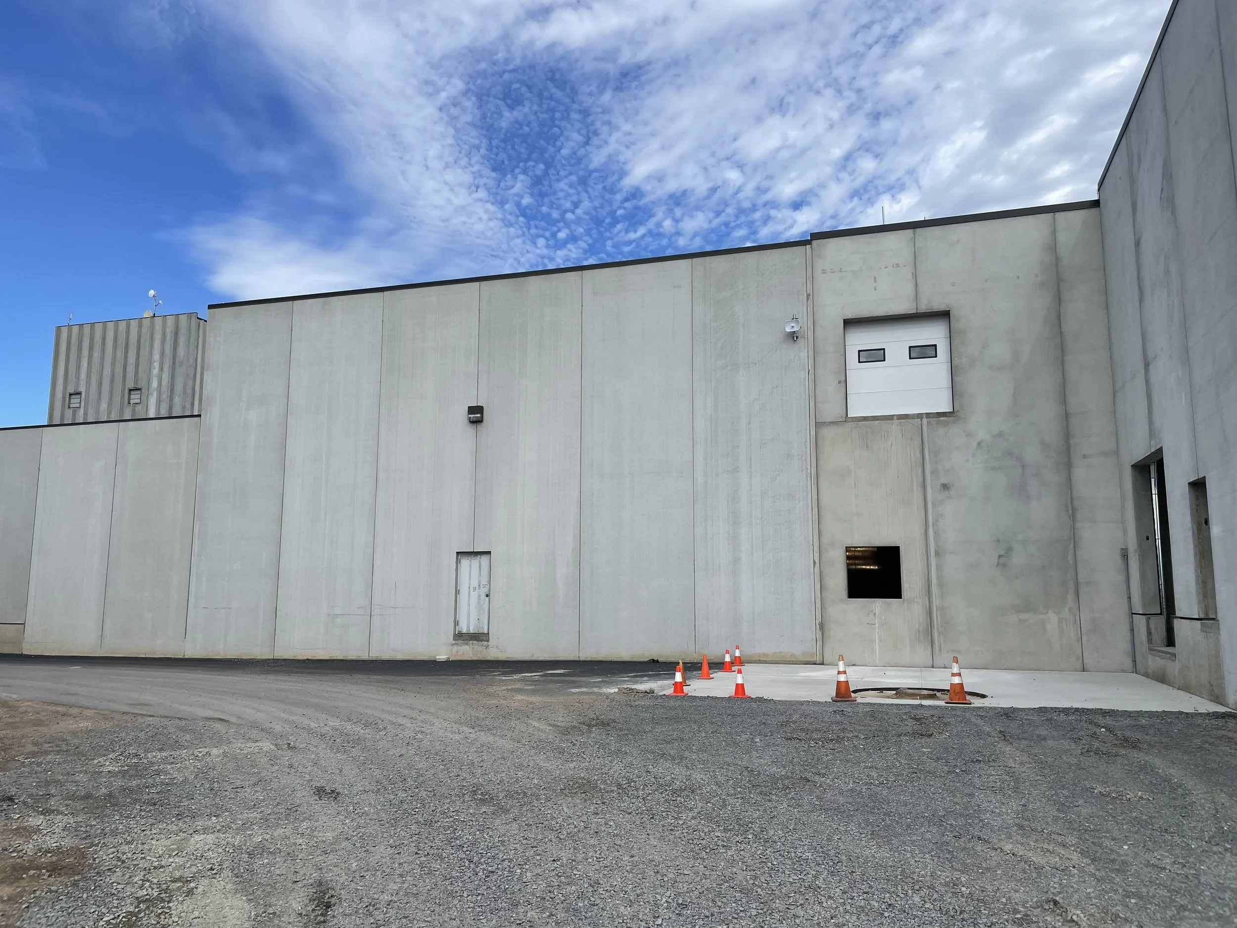 The image shows a large concrete building with minimal windows and a garage door. There are construction cones placed on the ground near an open area, and a cloudy blue sky overhead.
