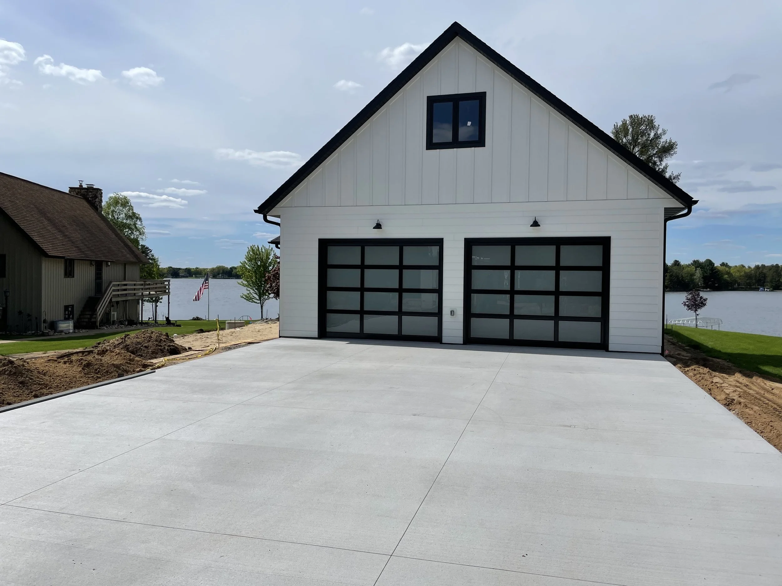A white modern garage with black-framed glass doors, a small window on the upper level, and exterior lights, situated by a lake with neighboring houses and a concrete driveway.