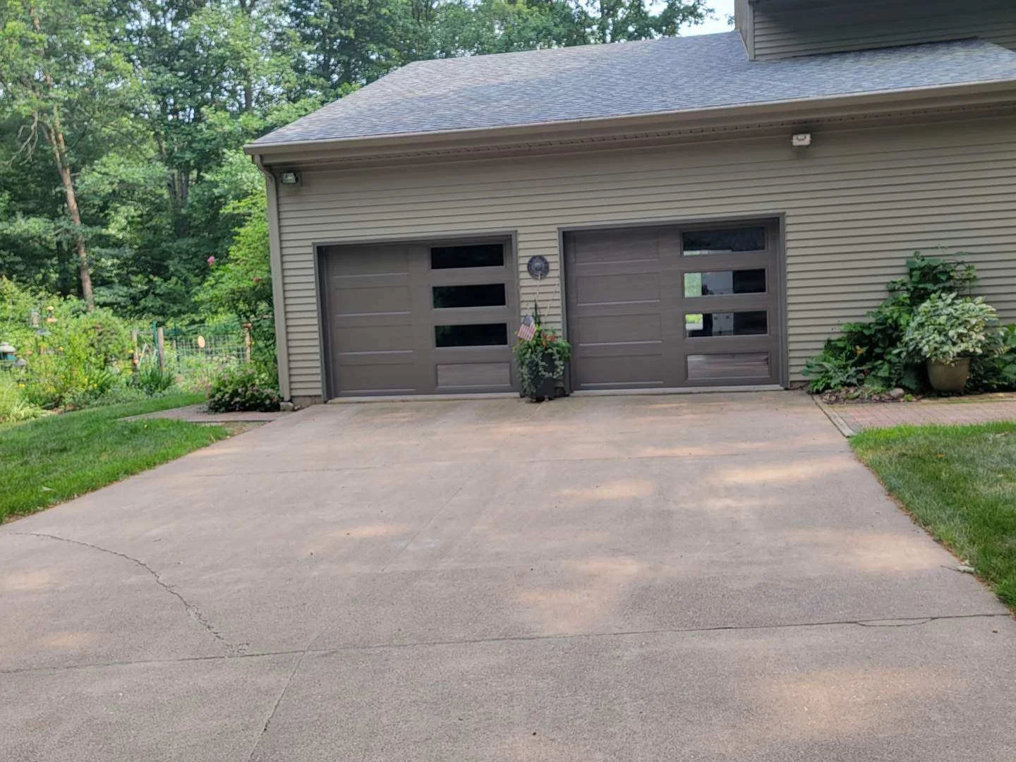 Two garage doors on a beige house with potted plants and a garden on either side, surrounded by trees and greenery.