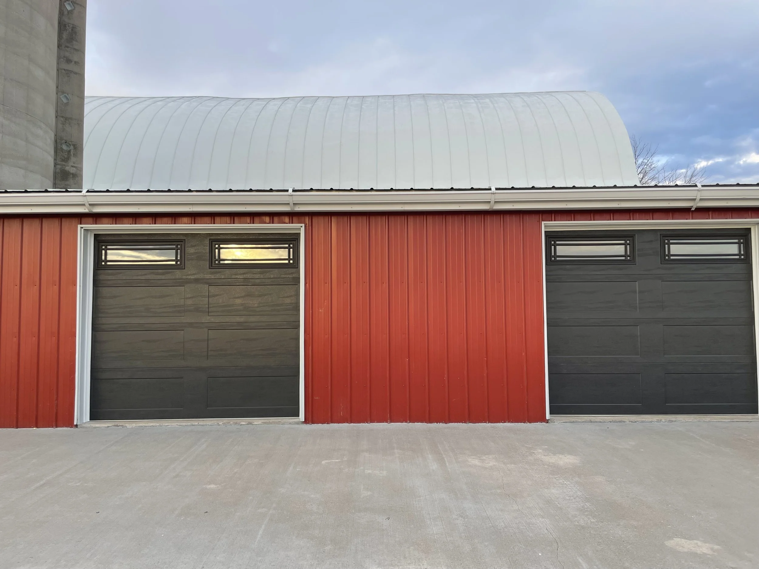 Modern garage with two black doors, red siding, metal roof, driveway, and concrete ground, under a blue sky with clouds.