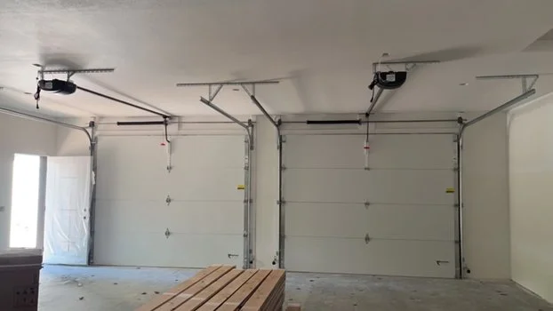 Empty garage with two roll-up doors, concrete floor, and garage door openers mounted on the ceiling.