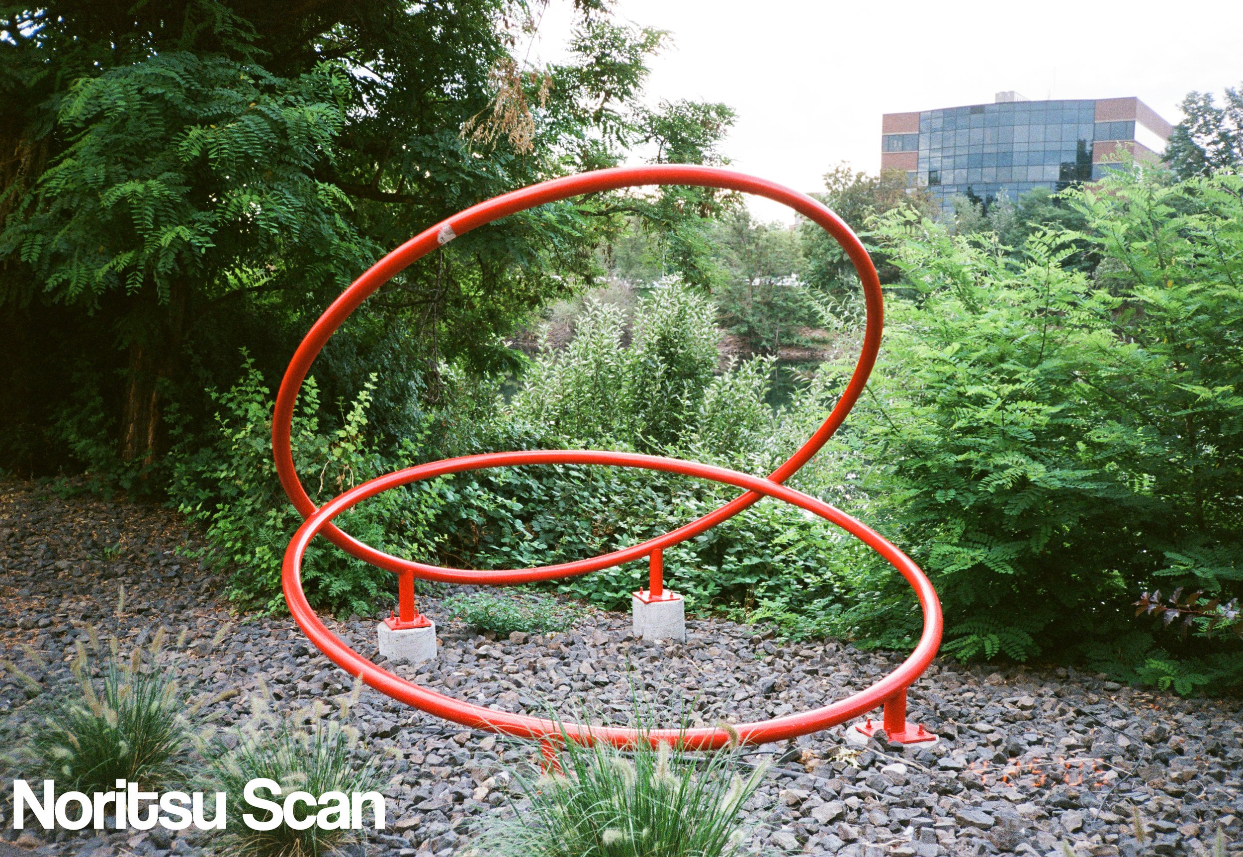 Contemporary outdoor sculpture with red circular and oval metal rings on concrete bases, situated on rocky ground amid lush green trees and bushes, with a modern glass office building in the background.