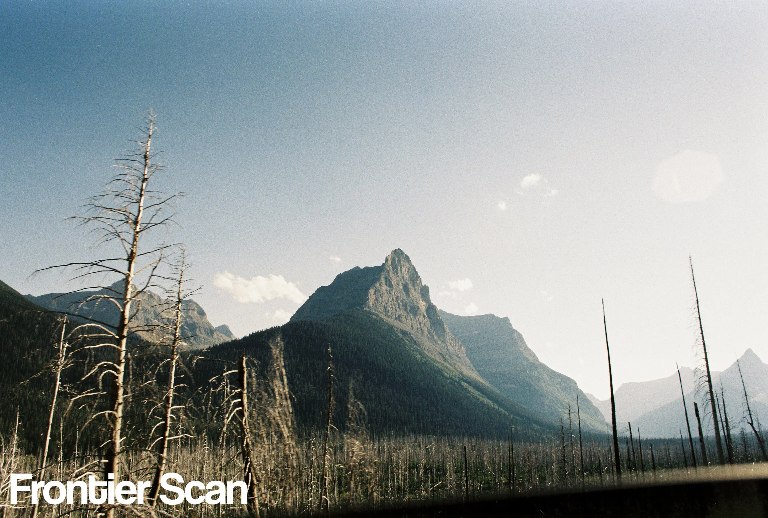 A mountain landscape with a large, jagged mountain peak in the background, surrounded by smaller mountains and a forested area in the foreground with several dead trees and a hazy sky.