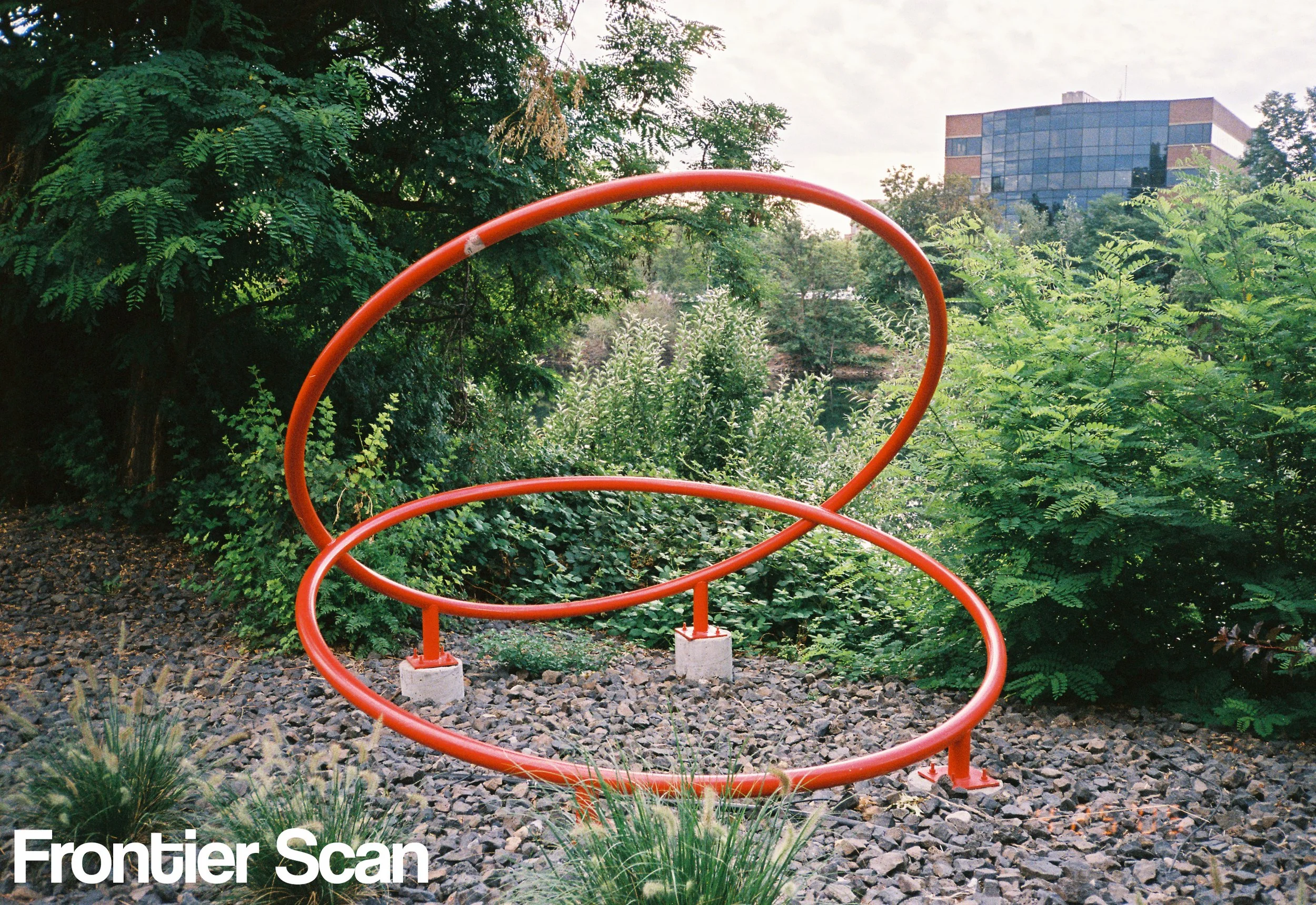 A modern orange sculpture with two large circular loops in a landscaped outdoor area surrounded by green foliage and a gravel ground, with an office building in the background.