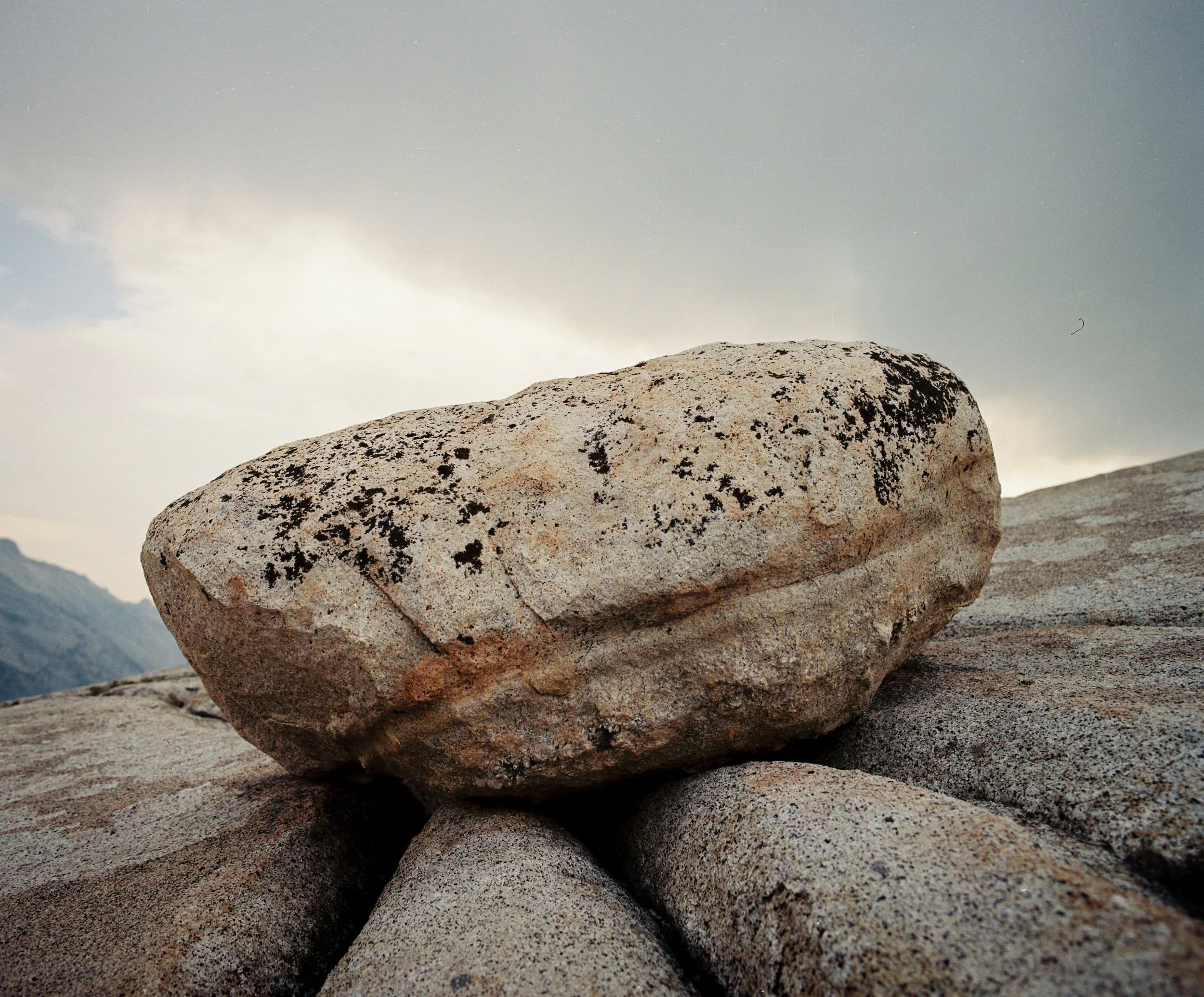 Close-up of a large, rounded rock with black lichen on its surface, resting on smaller rocks under a cloudy sky.