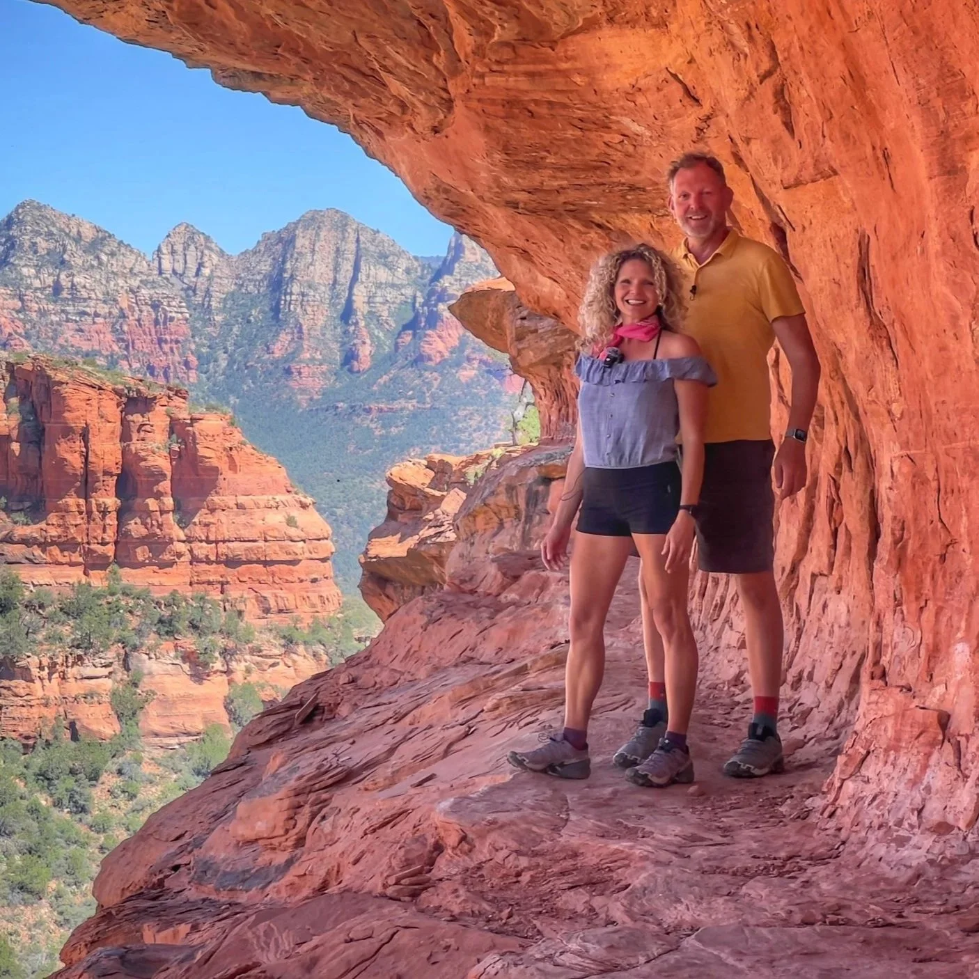 A man and a woman standing on a rocky ledge with a scenic desert canyon in the background, smiling at the camera.