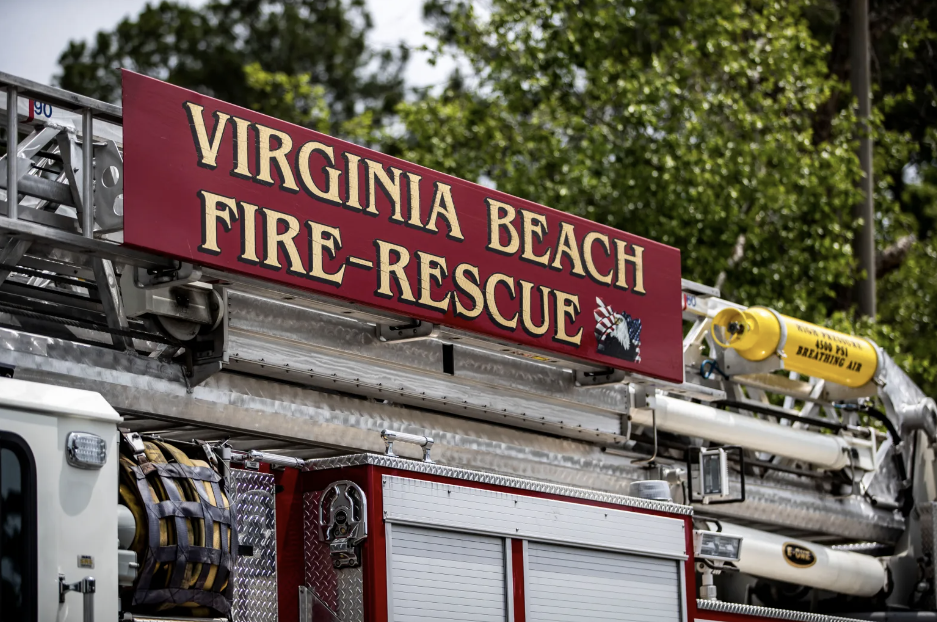 Close-up of a Virginia Beach fire rescue ladder truck with a red sign reading 'Virginia Beach Fire-Rescue' and a background of green trees.