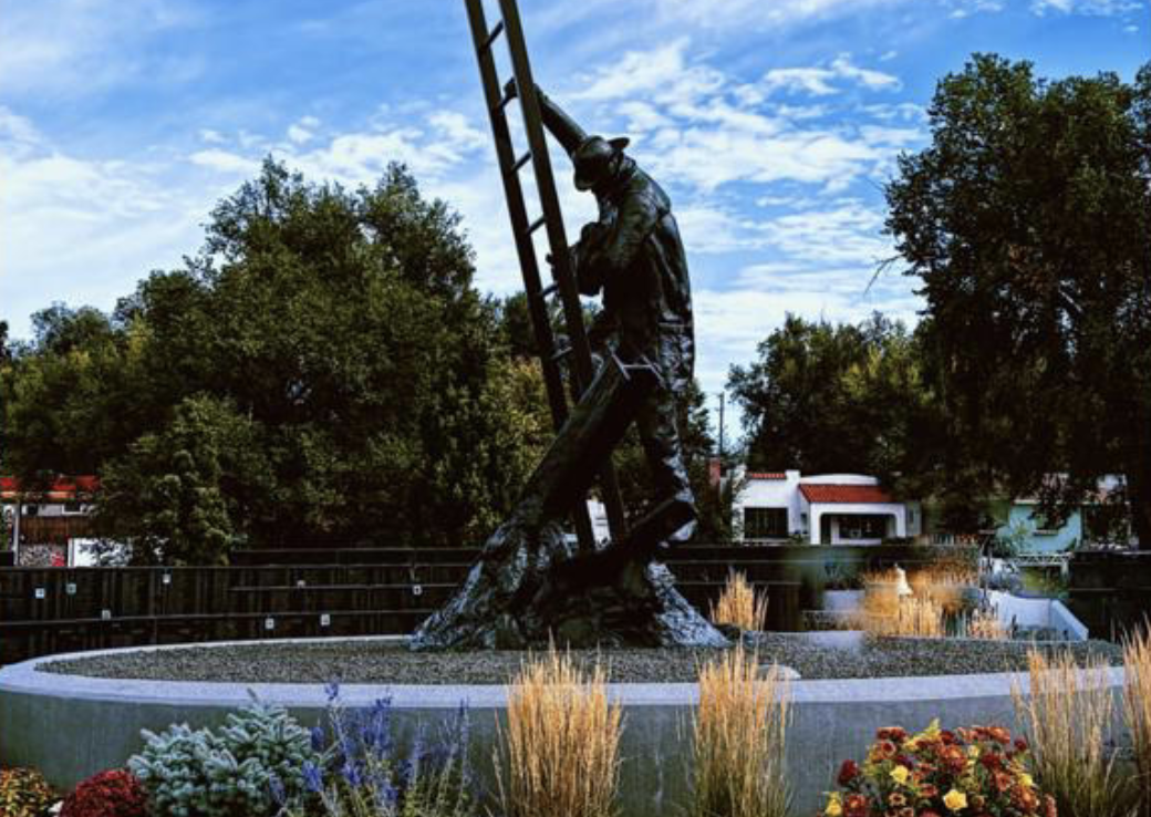 A large outdoor sculpture of a fireman climbing a ladder, situated in a garden with flowers and surrounded by trees, under a blue sky with scattered clouds.
