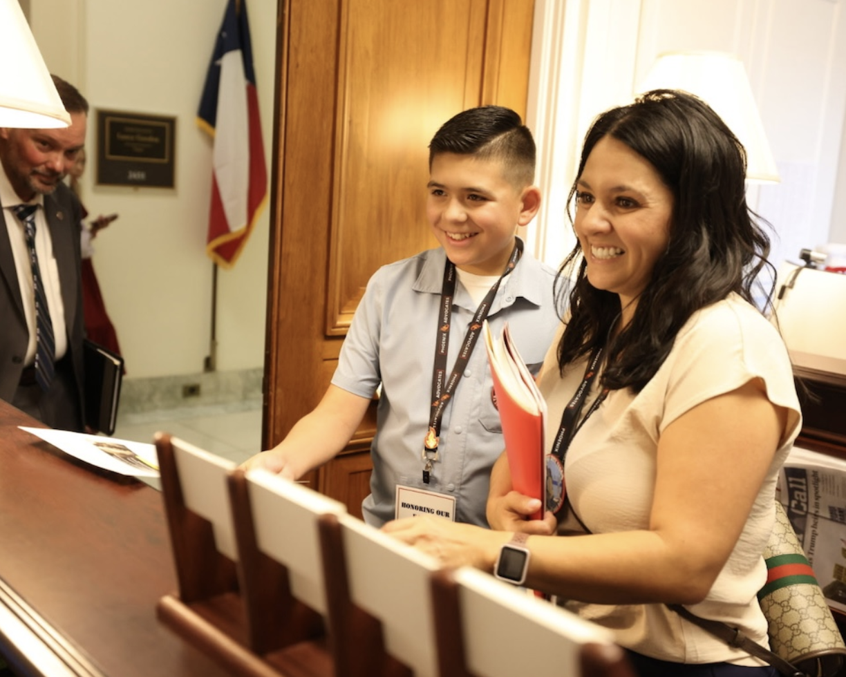A woman and a young boy smiling at a reception desk inside a building, with a man in a suit and a flag in the background.