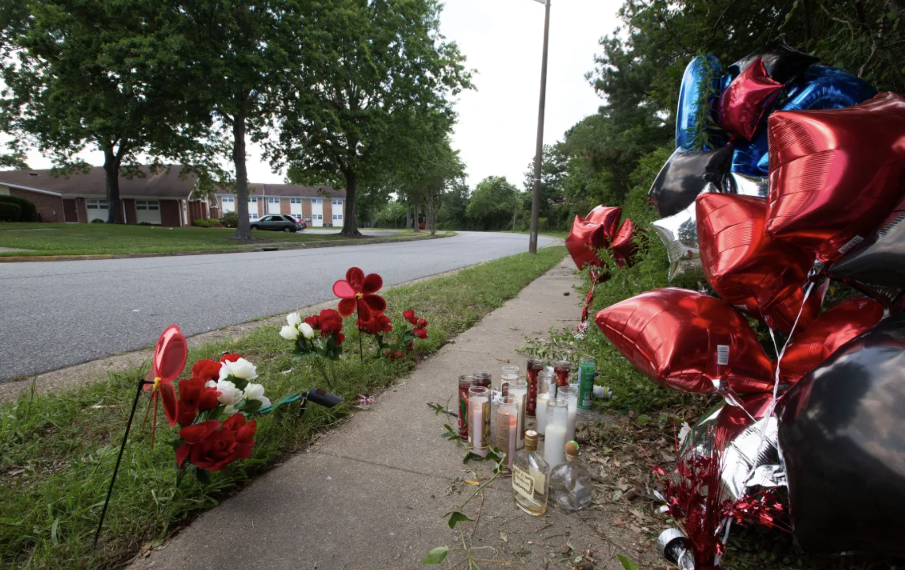 Memory site with flowers, candles, balloons, and tributes along a sidewalk in front of residential houses.