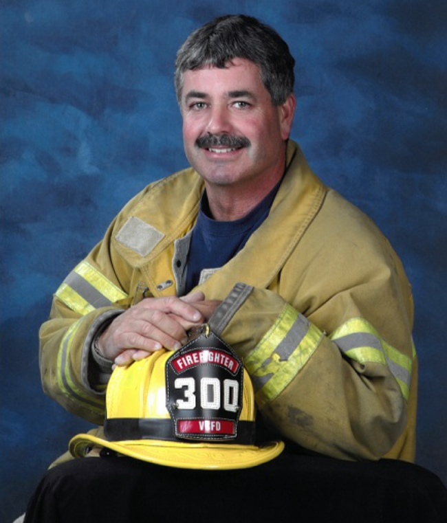 A firefighter sitting with a yellow helmet marked with the number 300 and the firefighter department badge, wearing a tan uniform with reflective stripes, in front of a blue textured background.