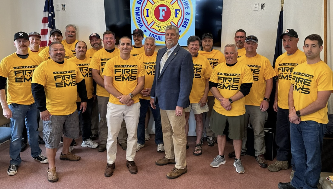 Group of men, some wearing yellow T-shirts with 'FIRE EMS' printed on them, standing with a man in a blue blazer in front of a Virginia Fire Fighters and EMTs logo during a formal event.