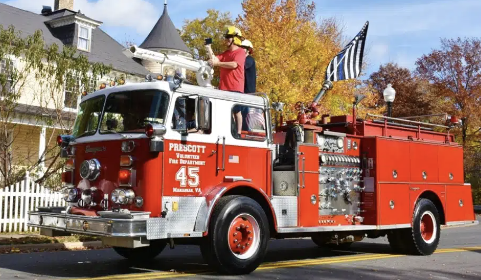 A red fire truck with the number 45 and the words Prescott Volunteer Fire Department on the side, driving down a street with autumn trees in the background. 