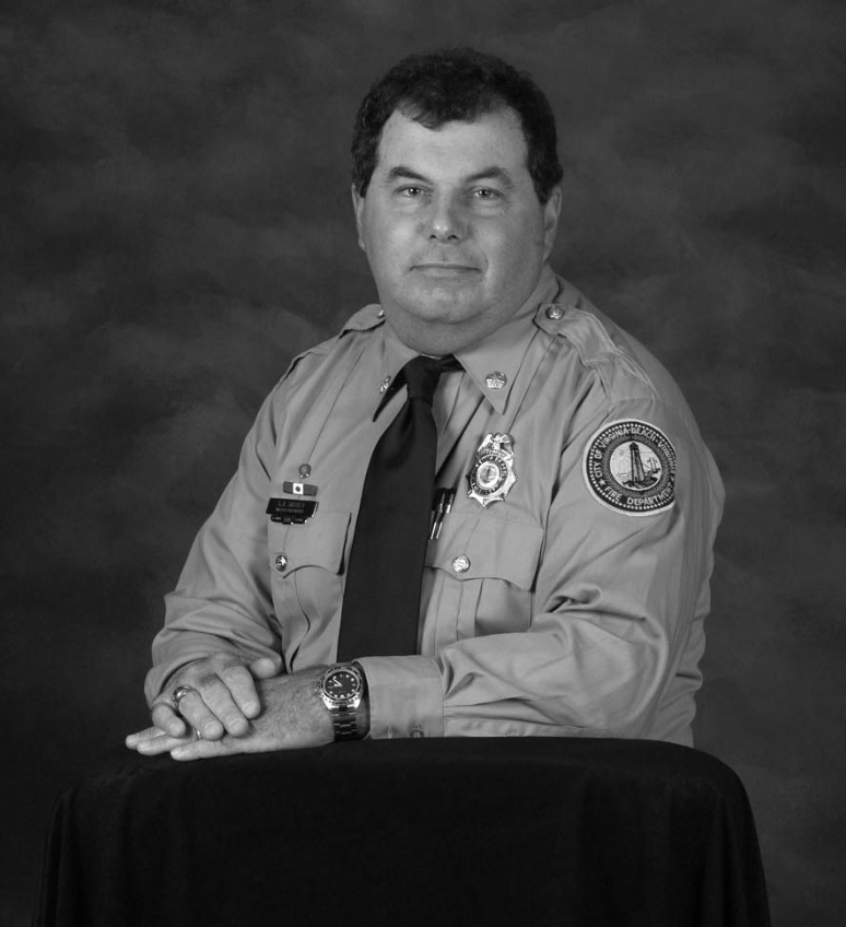 Black and white portrait of a male firefighter in uniform, sitting at a table with his hands crossed, facing the camera.