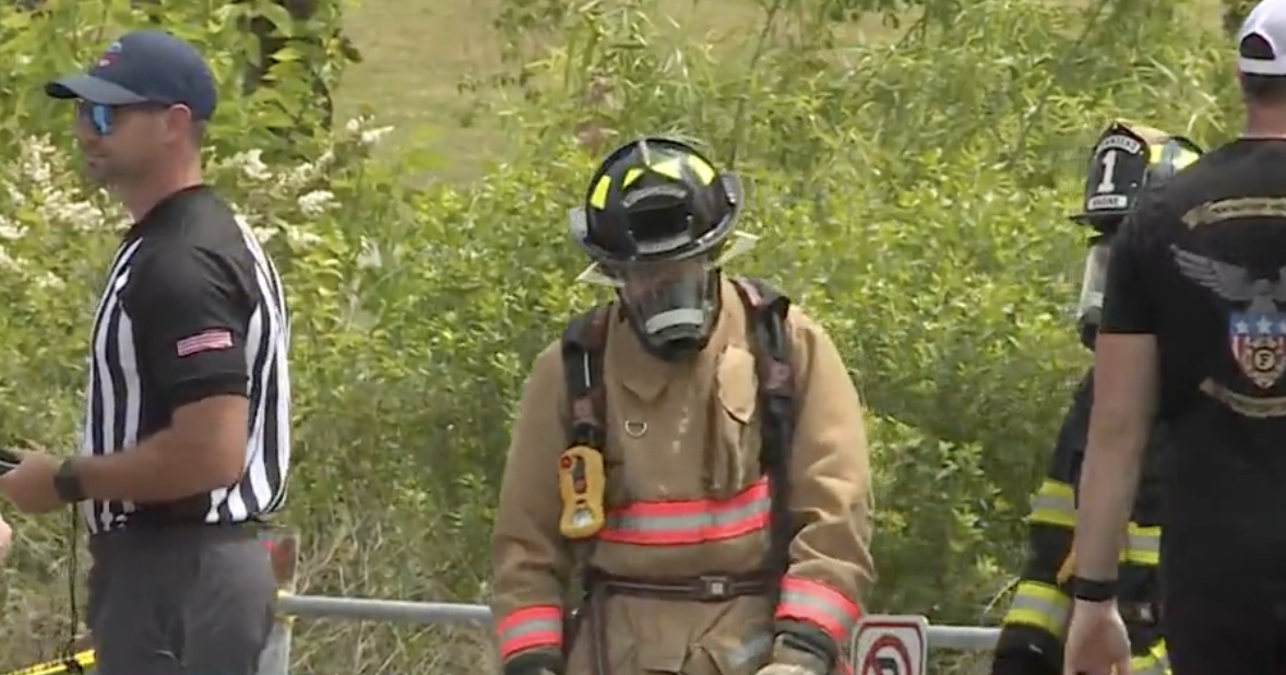 Firefighter with protective gear and helmet standing outdoors, surrounded by greenery, along with other individuals.