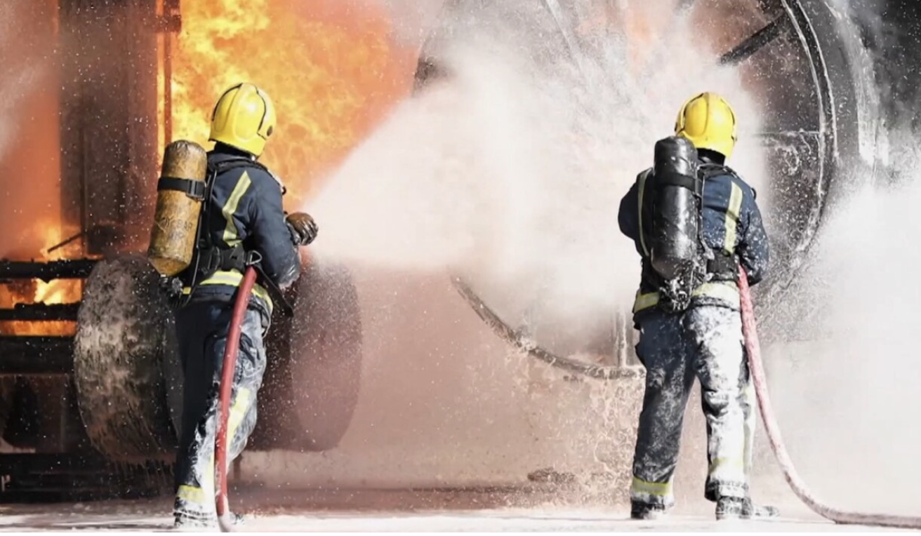 Two firefighters in protective gear and yellow helmets are spraying water on a large fire inside a building.