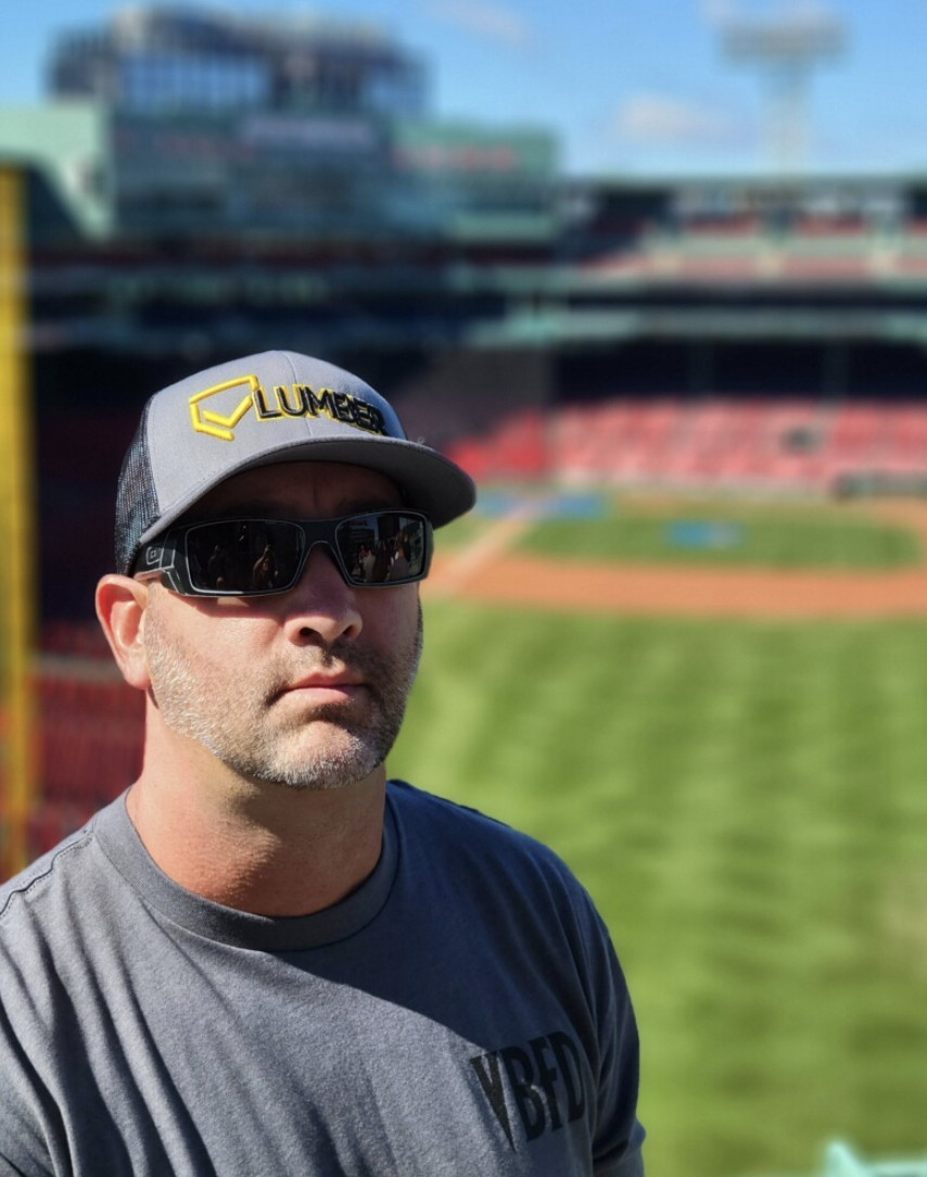 A man wearing sunglasses, a gray cap with a yellow logo, and a gray T-shirt with black text, with a stadium in the background.