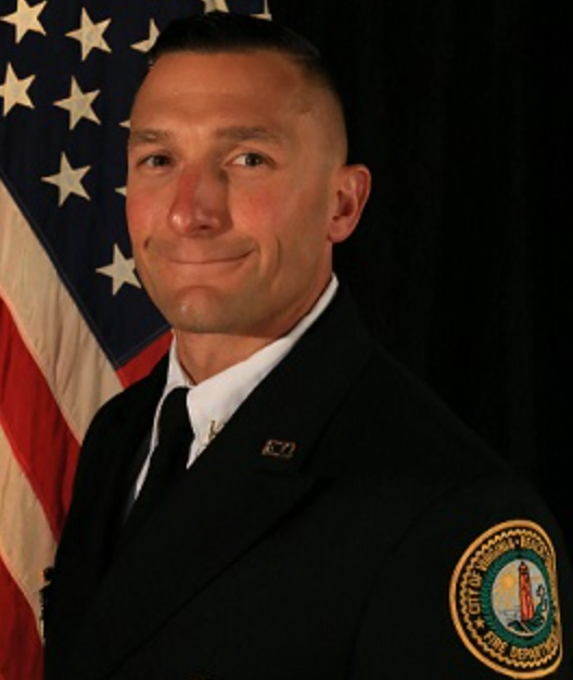 A man in formal uniform standing in front of an American flag.